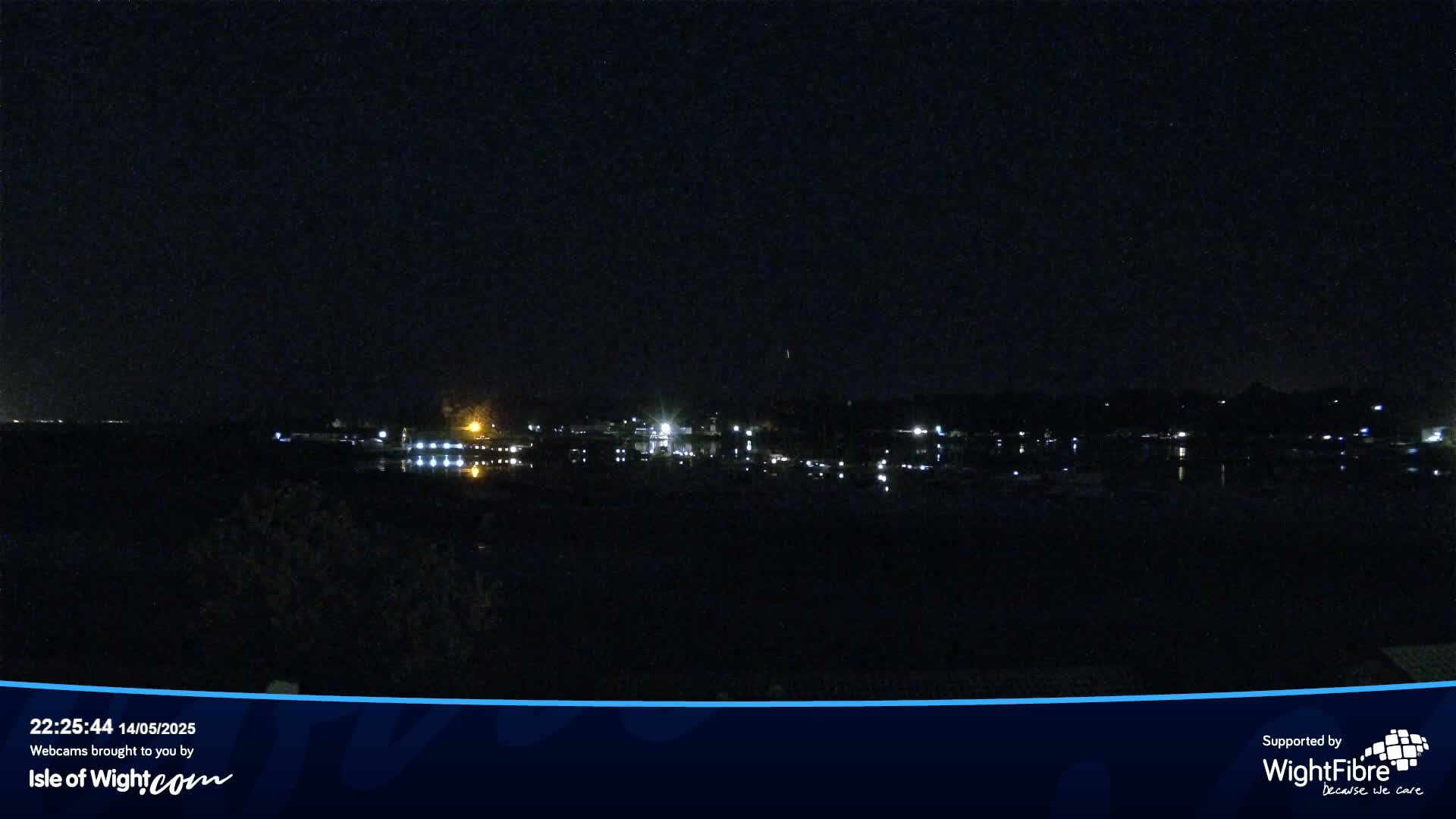 A nighttime view of a calm body of water reflecting the lights of a distant town under a clear, dark sky.