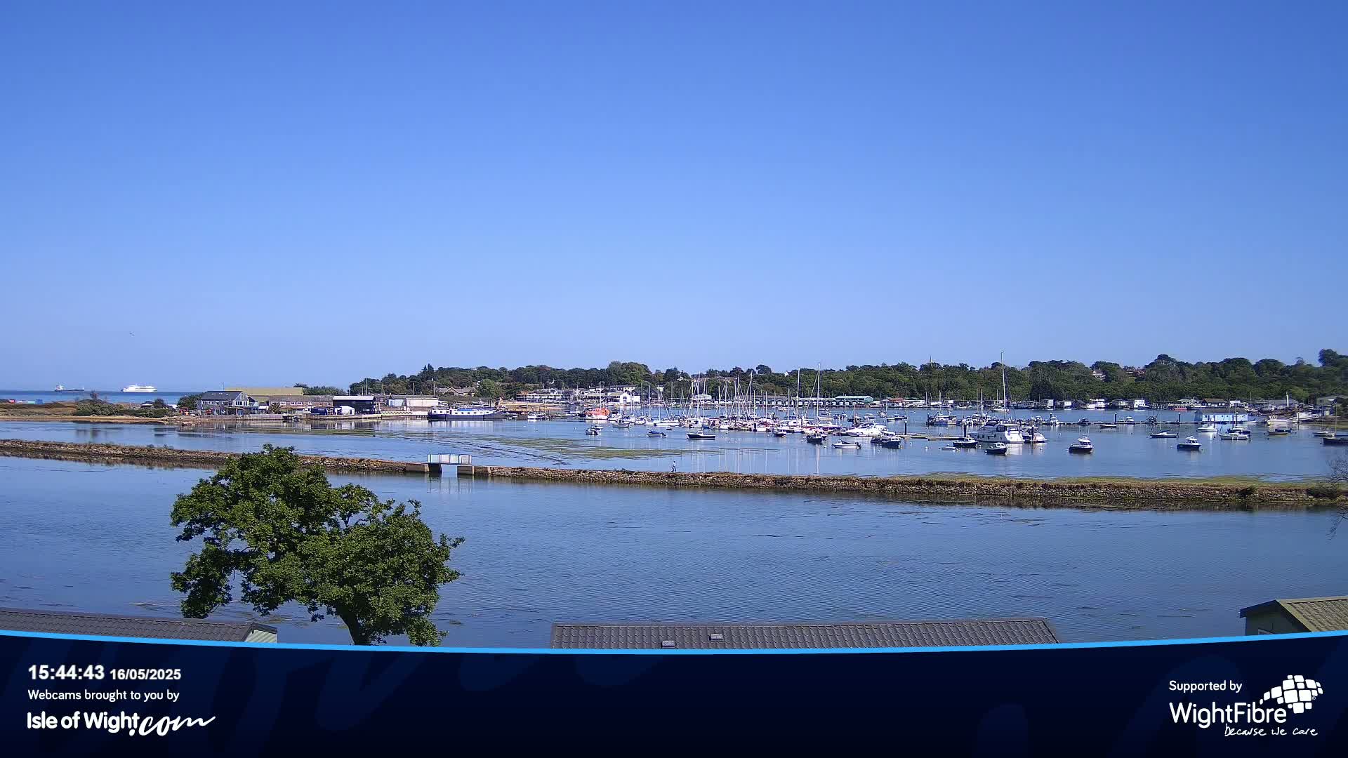 A calm, sunny day shows a marina filled with numerous boats and yachts, situated in a bay near a tree-lined shoreline.