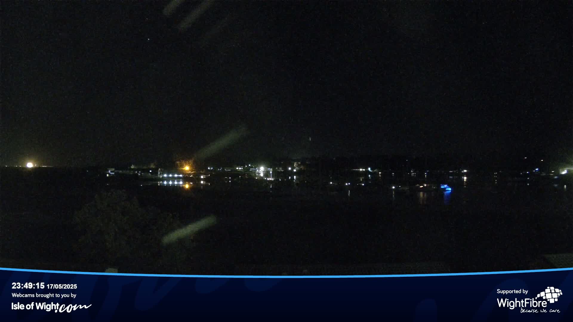 A nighttime view of a calm body of water with dimly lit buildings and boats on the far shore under a clear, starry sky.
