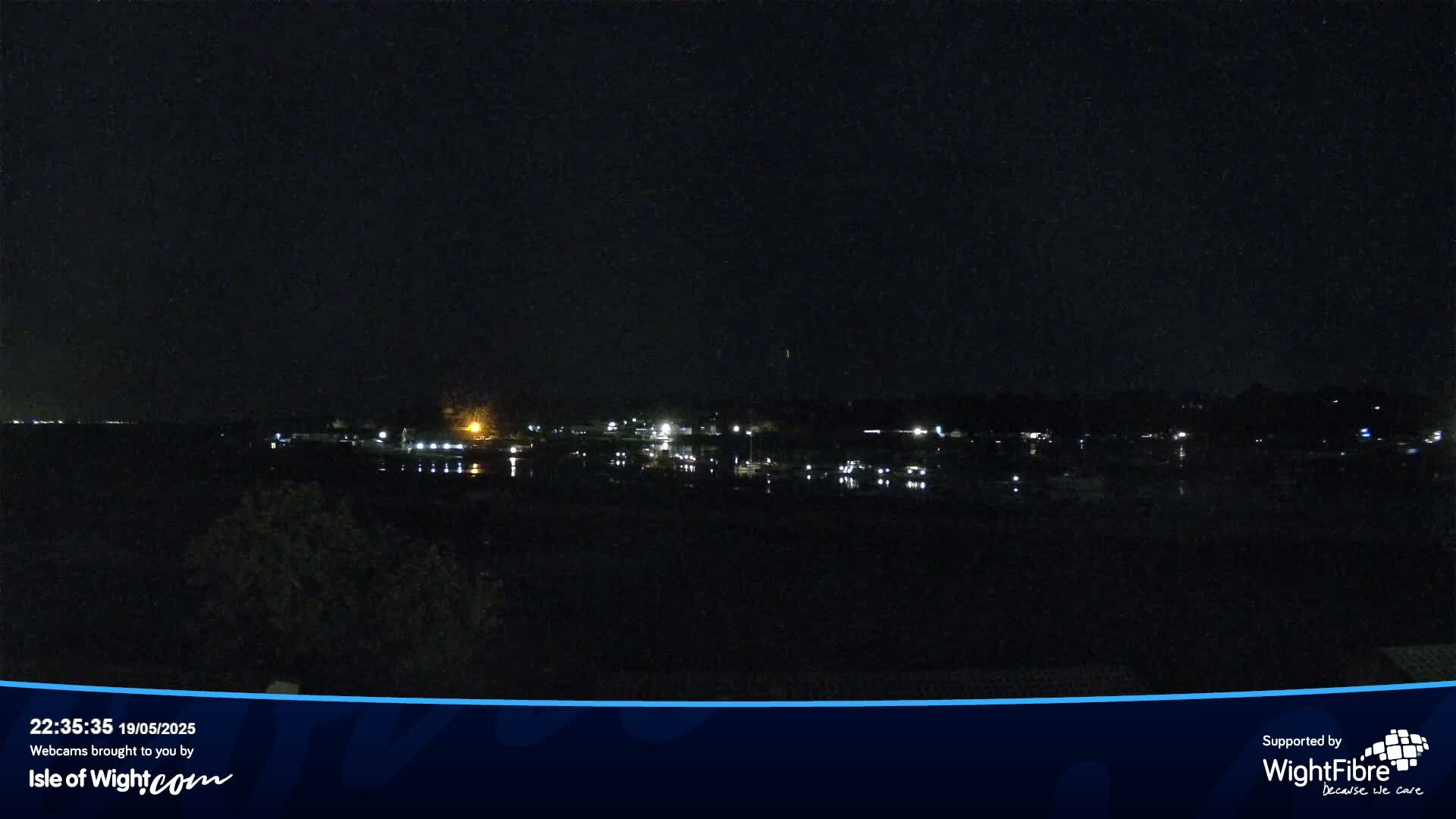 A nighttime view of a dark body of water with dimly lit buildings and boats across the shore under a clear, dark sky.