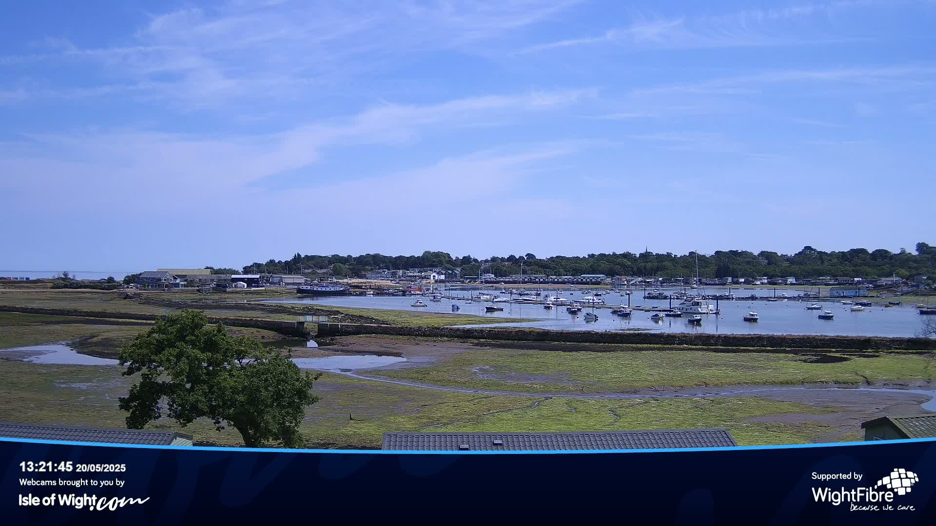 A sunny day reveals a harbor filled with boats, nestled beside a shoreline of exposed mudflats and greenery, with a village visible in the background.
