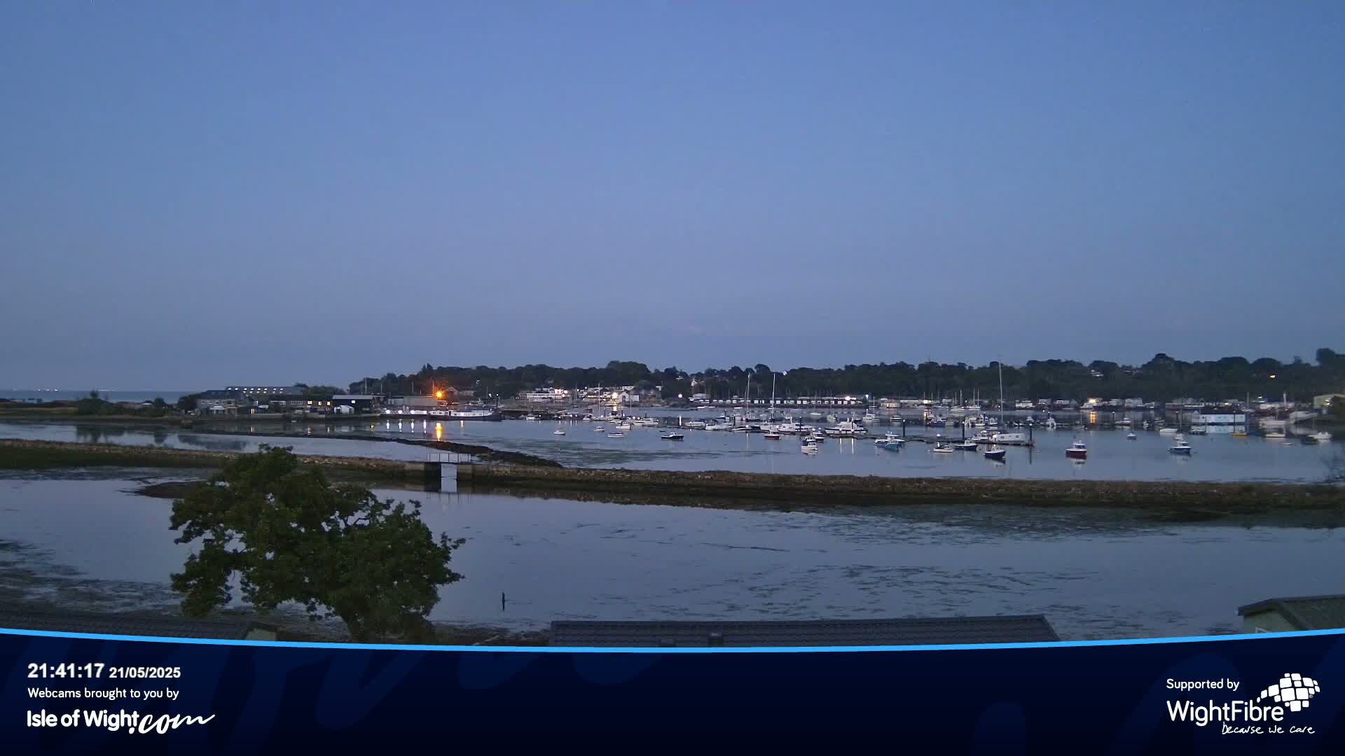 A calm, twilight scene shows a harbor filled with boats, nestled beside a shoreline with a few trees and buildings under a clear, dusky sky.