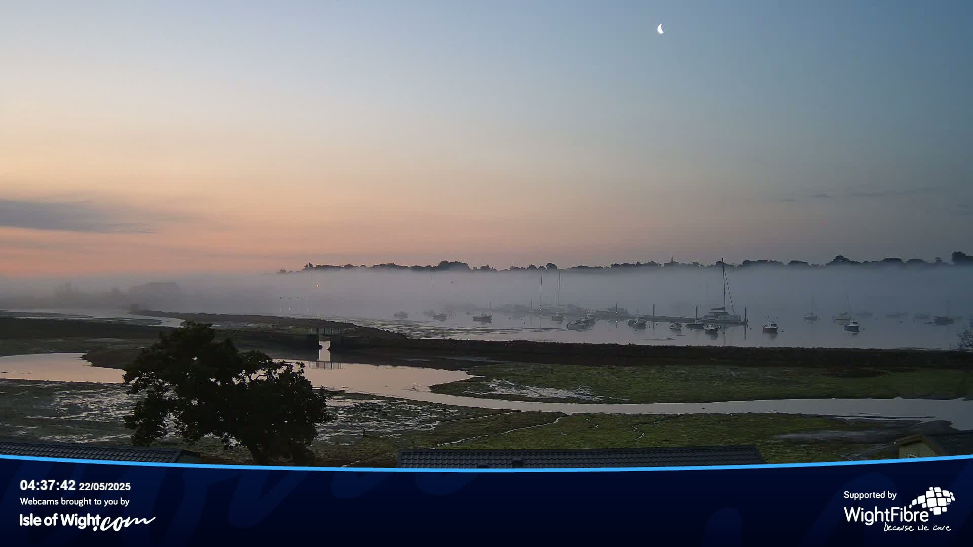 A foggy sunrise over a calm body of water with several boats docked, a crescent moon visible in the pale sky.