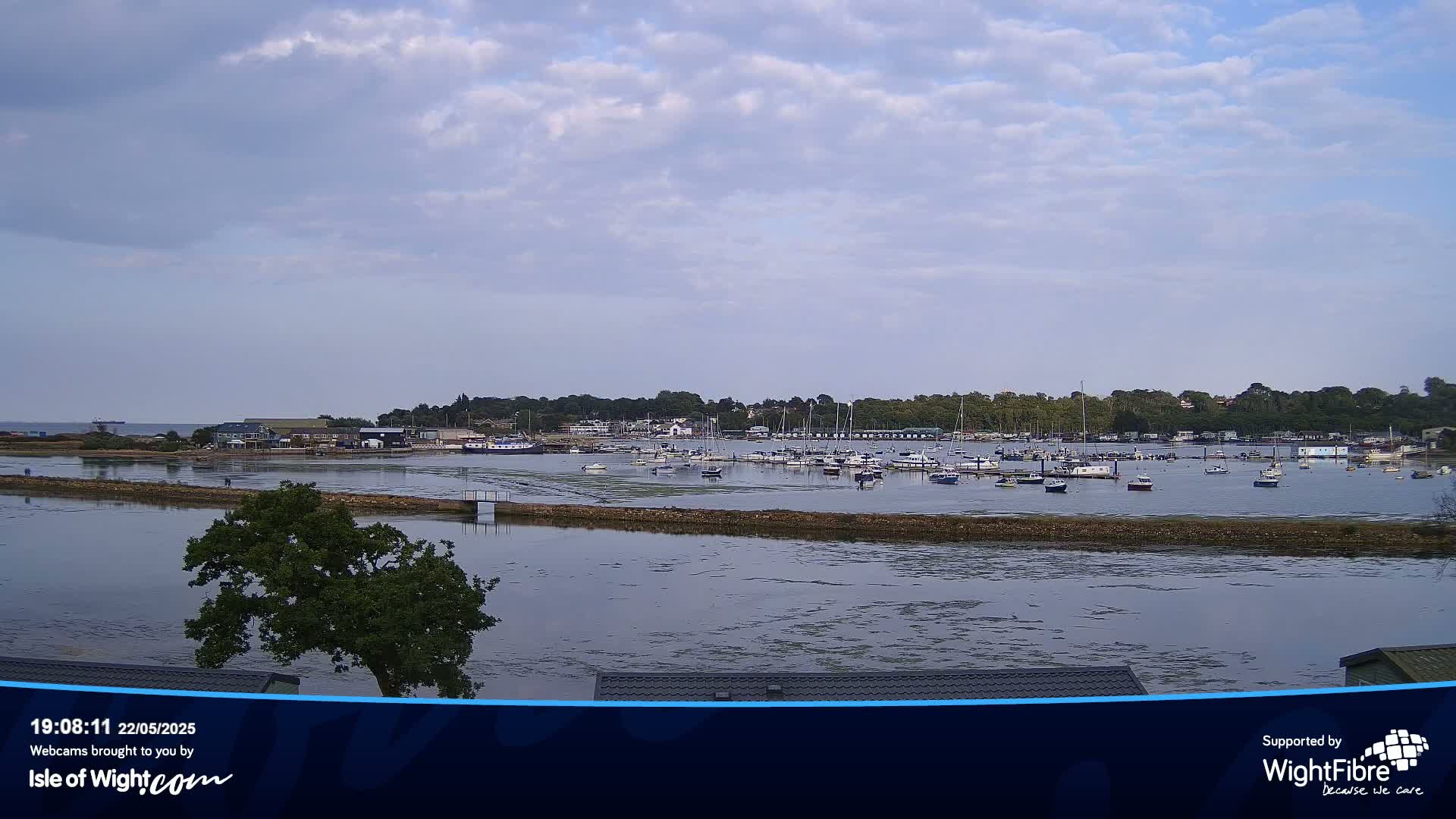 A calm bay with numerous boats moored, under a partly cloudy sky, is viewed from a slightly elevated position.