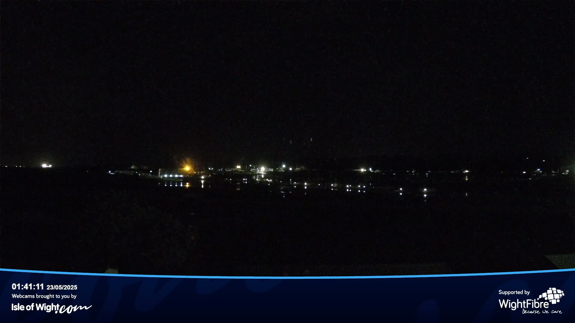 A nighttime view of a dark body of water reflecting the lights of a distant town under a clear, dark sky.
