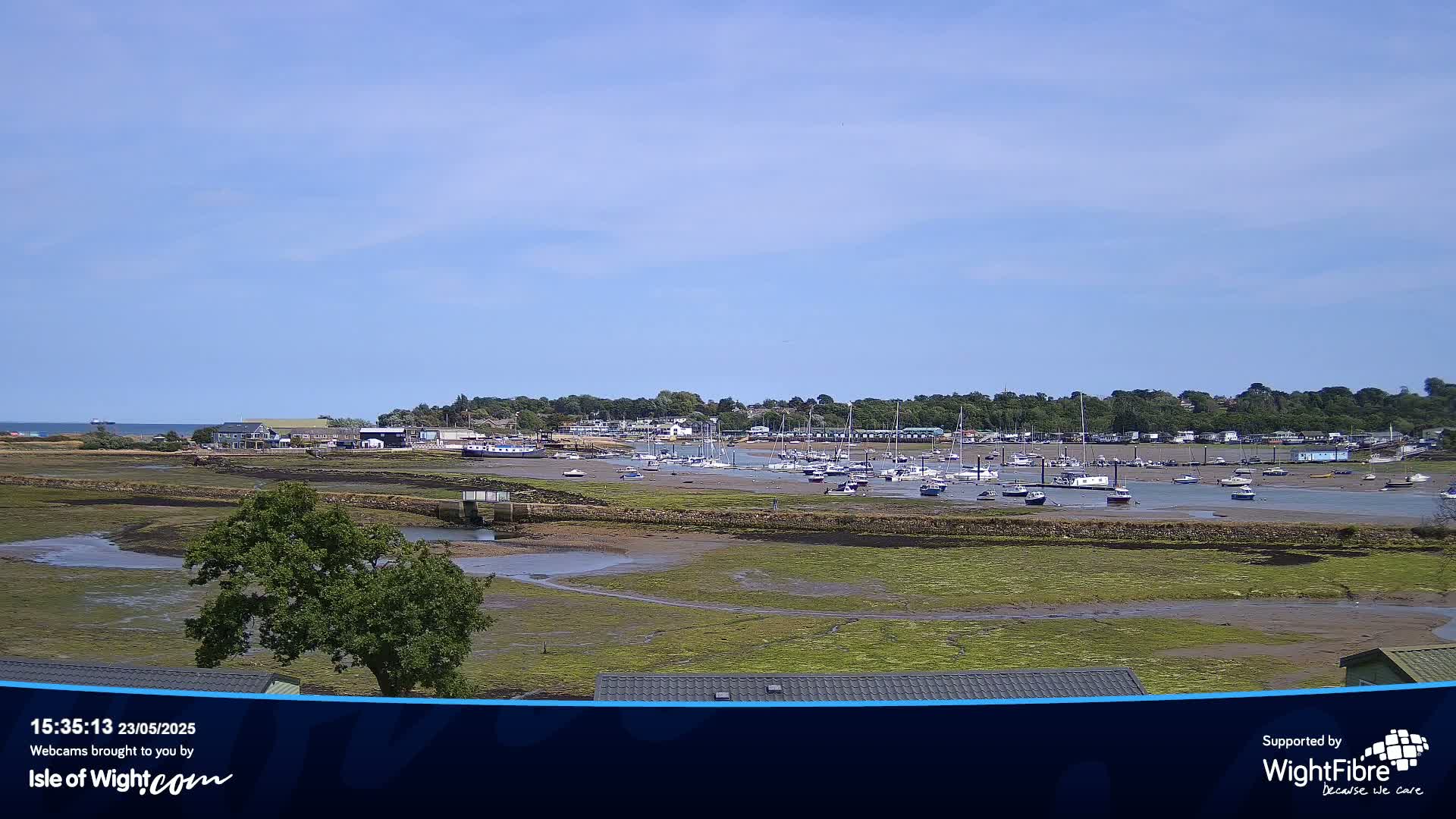 A mostly sunny day reveals a harbor filled with numerous boats at low tide, surrounded by exposed mudflats and a distant tree-lined shoreline.