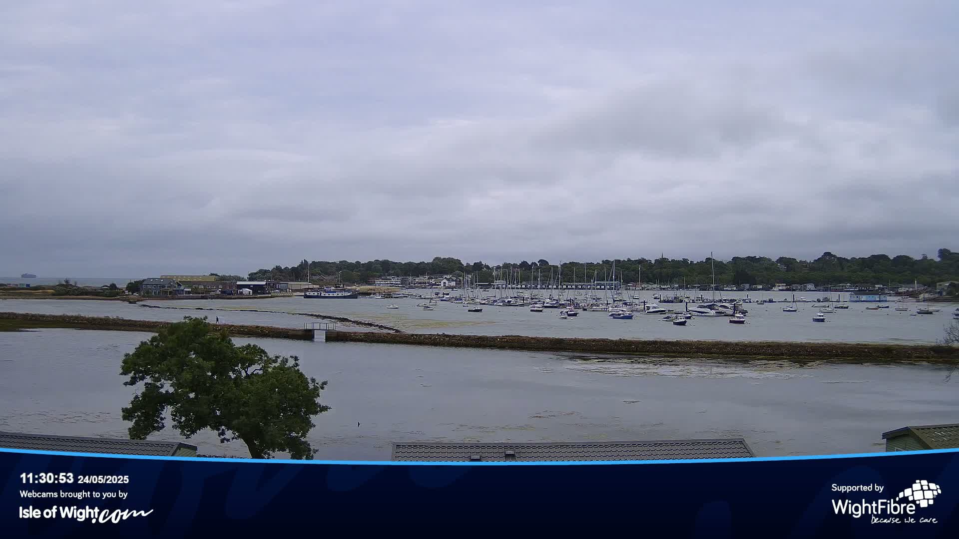 A calm, overcast day shows a harbor filled with numerous boats, partially sheltered by a landmass with trees and buildings.