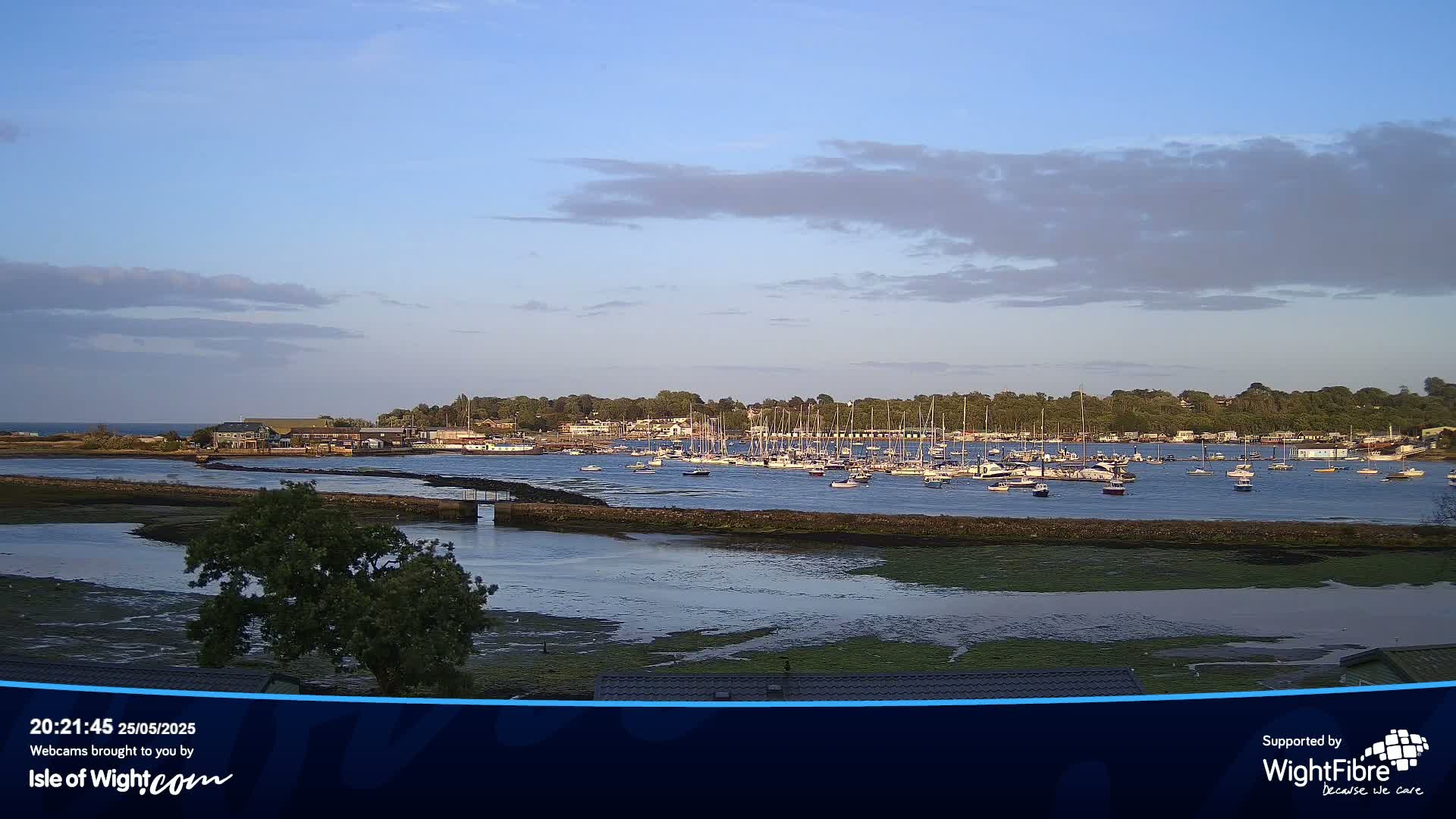 A calm body of water with numerous boats at anchor is nestled between low-lying land, under a partly cloudy sky.