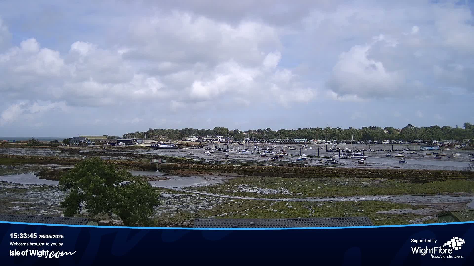 A mostly cloudy day reveals a harbor with many boats at low tide, surrounded by green land.