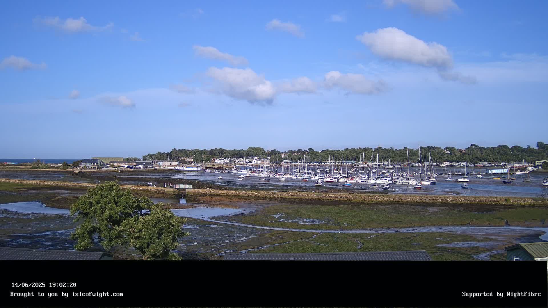 A harbor filled with numerous sailboats and motorboats is visible at low tide under a mostly sunny sky with scattered clouds.