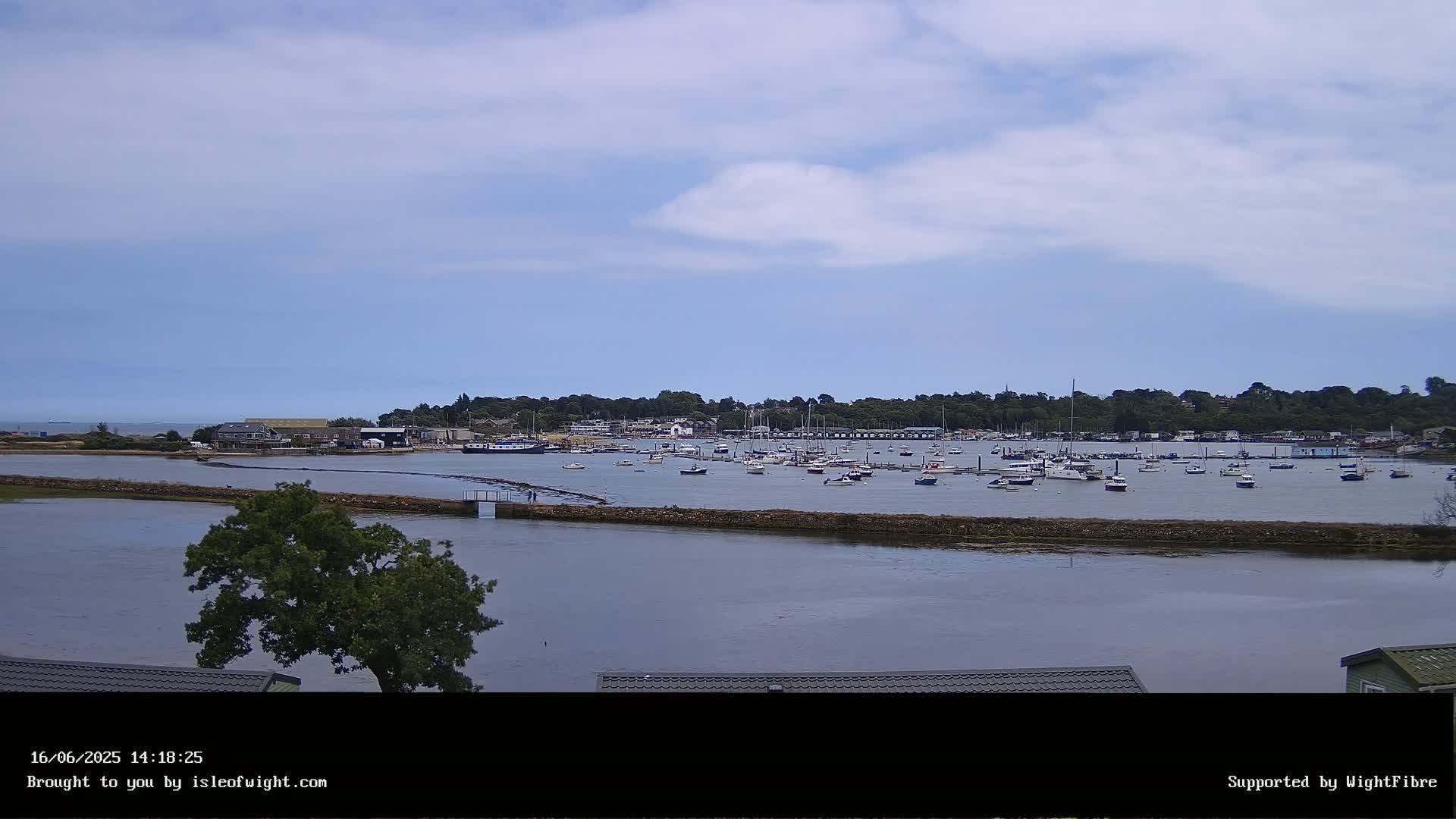 A calm, partly cloudy day shows a harbor filled with numerous boats and yachts, surrounded by a green shoreline.