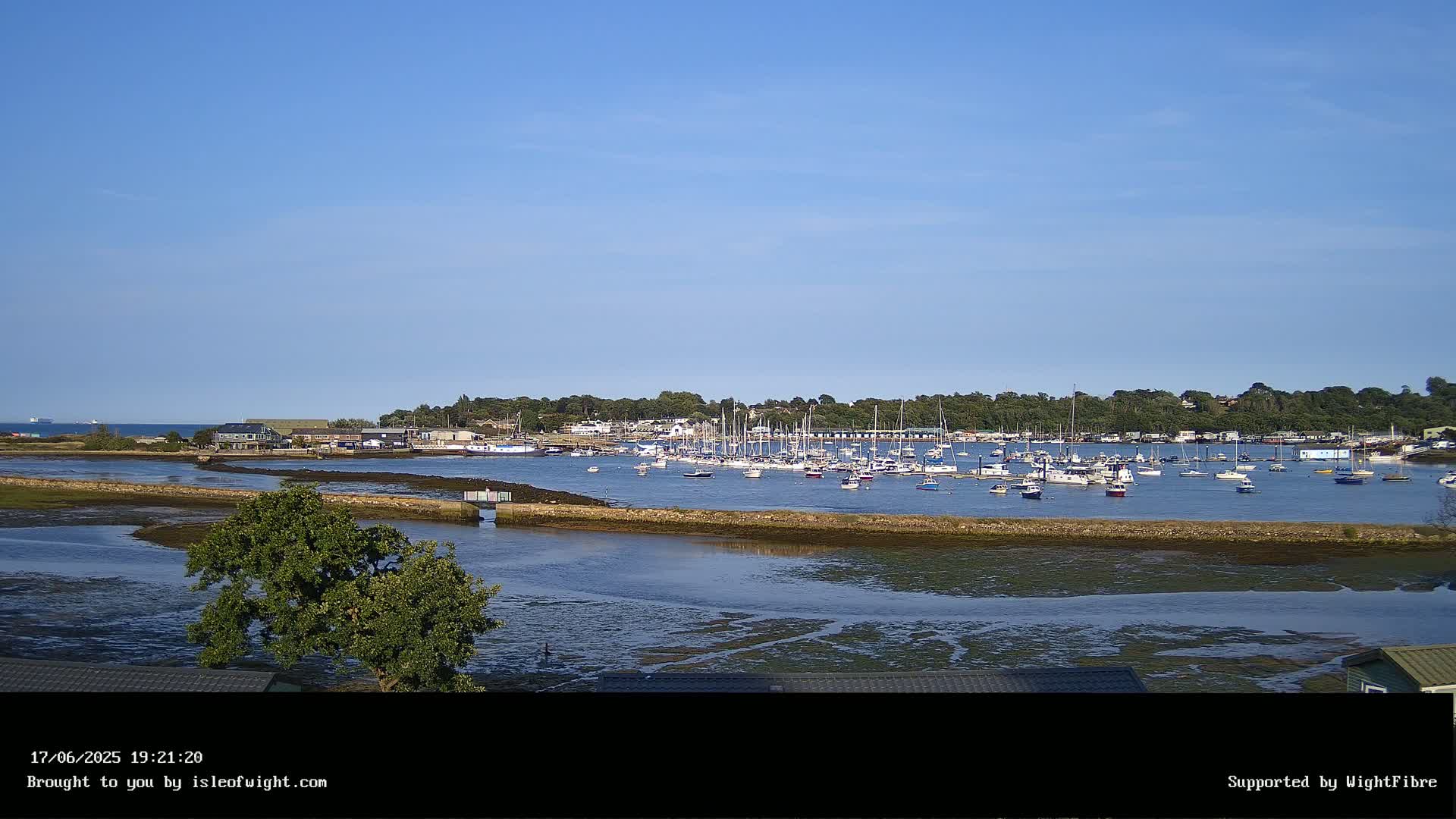 A calm, sunny day reveals a harbor filled with numerous sailboats and motorboats, nestled beside a tree-lined shore.