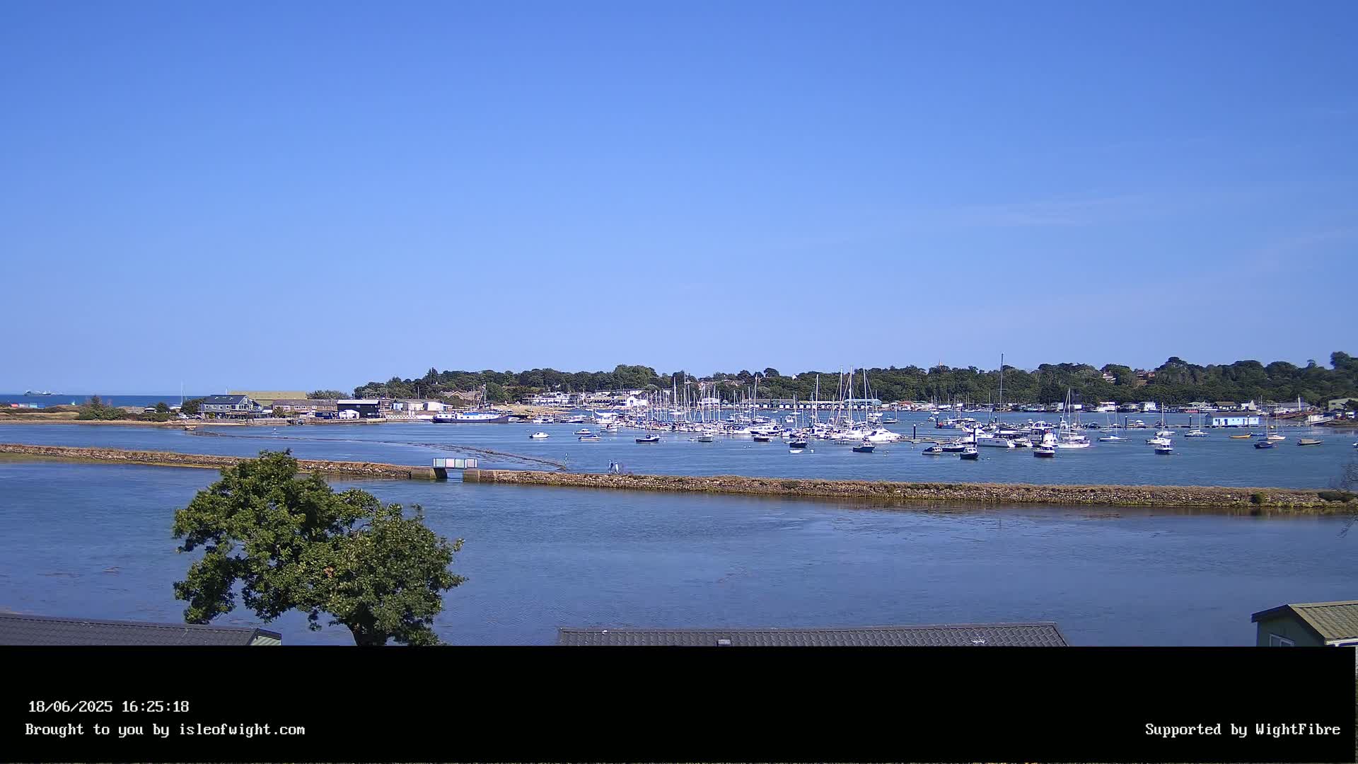 A calm, sunny day reveals a harbor filled with numerous sailboats and motorboats, nestled behind a low-lying landmass with trees and buildings, under a clear blue sky.