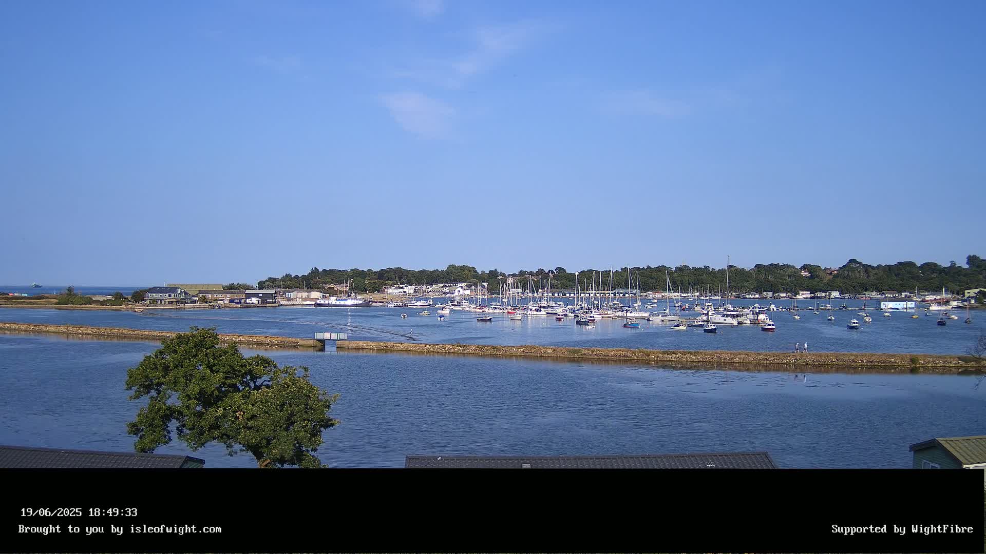 A calm, sunny day reveals a harbor filled with numerous sailboats and motorboats, nestled beside a verdant shoreline.