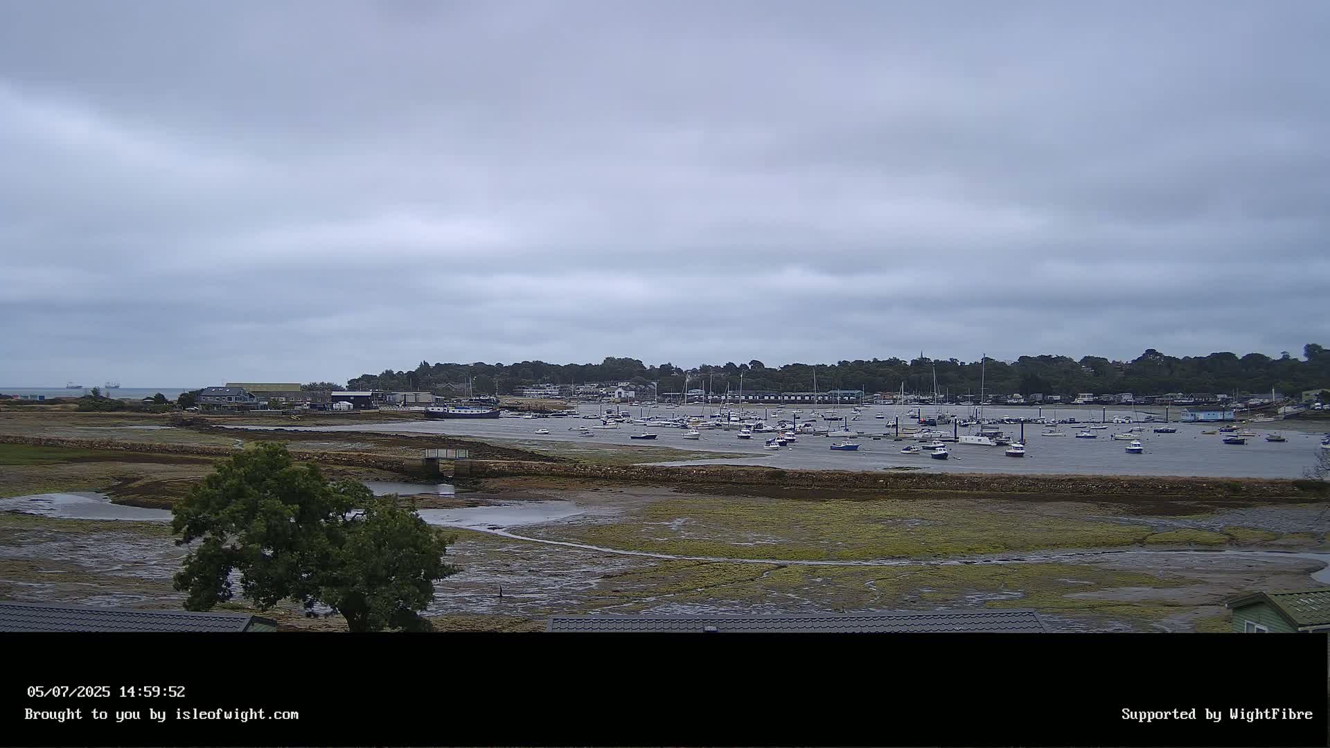 A mostly cloudy day reveals a harbor filled with numerous boats at low tide, backed by a tree-lined shoreline with buildings.
