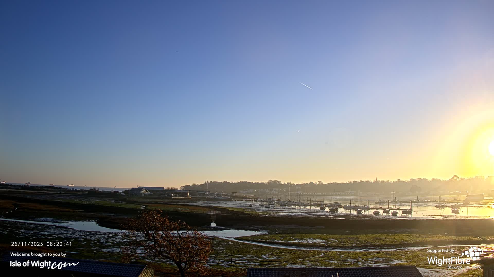 A clear and sunny morning illuminates a vast tidal estuary filled with moored boats on exposed mudflats and winding water channels, with a distant treeline and buildings beneath a bright low sun.