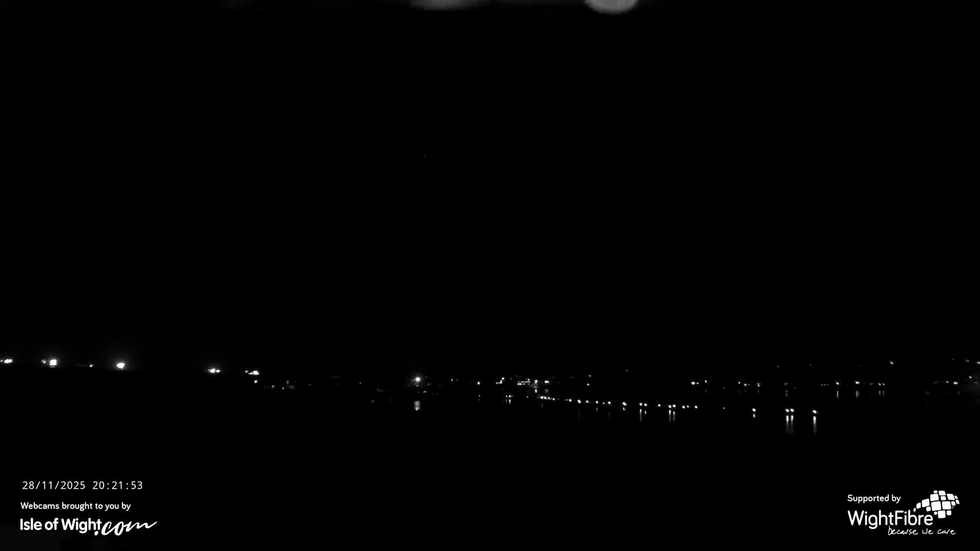 The image displays a very dark nighttime scene with numerous distant lights from a shoreline or urban area reflecting on calm water, under what appears to be a clear sky.