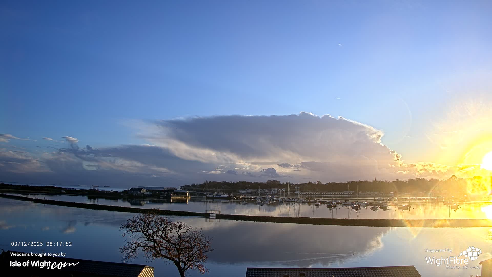 A vibrant sunrise illuminates a tranquil harbor, where numerous boats are moored on calm, reflective waters, bordered by a treelined shore, beneath a partly cloudy sky dominated by a large, dramatic cloud formation.