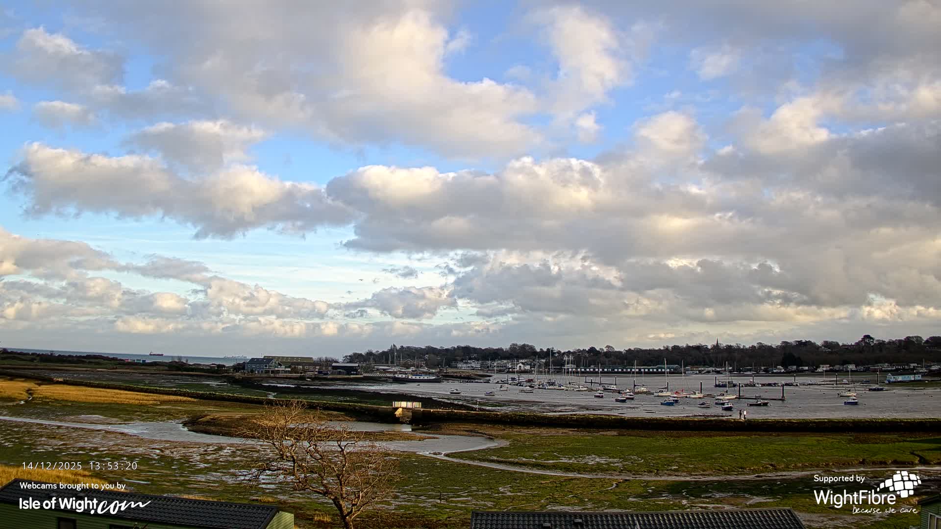 A vibrant sunrise illuminates a tranquil harbor, where numerous boats are moored on calm, reflective waters, bordered by a treelined shore, beneath a partly cloudy sky dominated by a large, dramatic cloud formation.