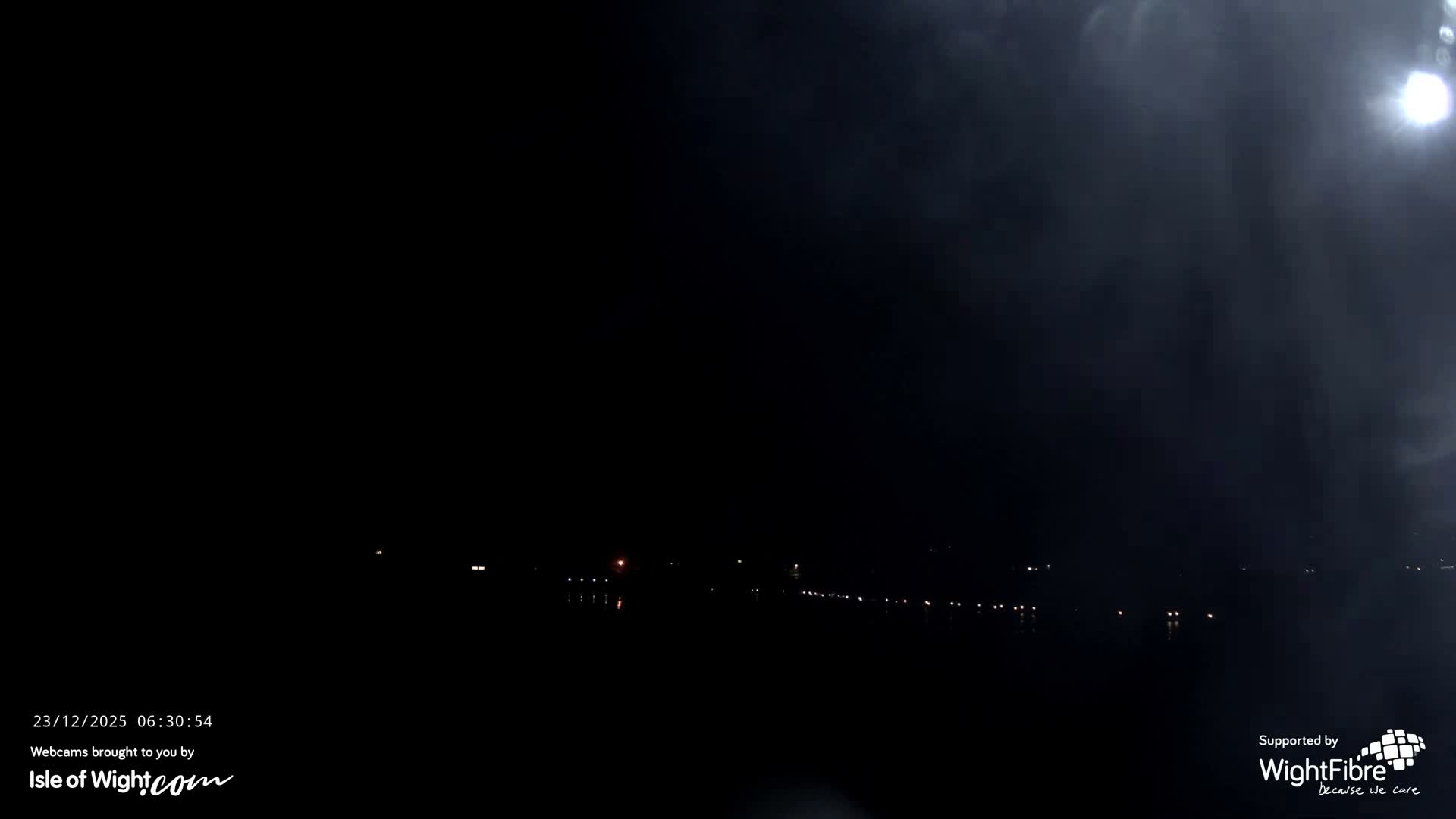 The image depicts a dark, cloudy night over what appears to be a body of water, with a line of distant lights visible across the horizon and a bright, diffused light source in the upper right sky.