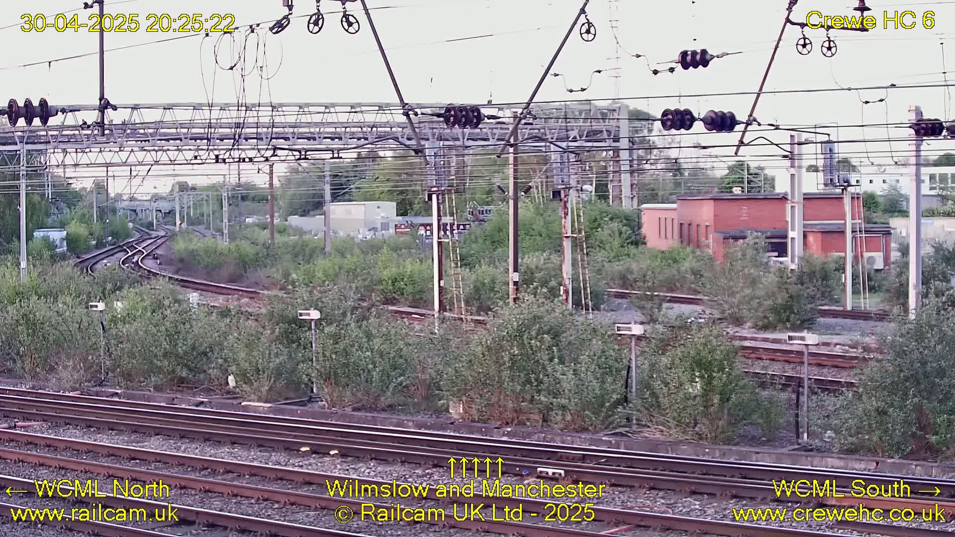 A view of railway tracks curving through overgrown vegetation under an overcast sky, with overhead electrical lines and a brick building in the background.