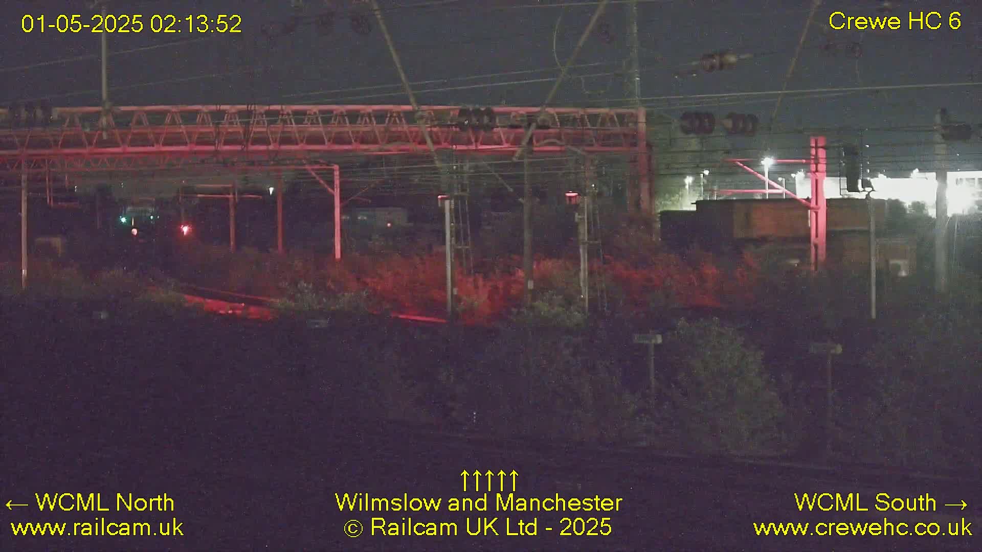 A nighttime view of a railway line under a metal structure, with dimly lit vegetation on either side.