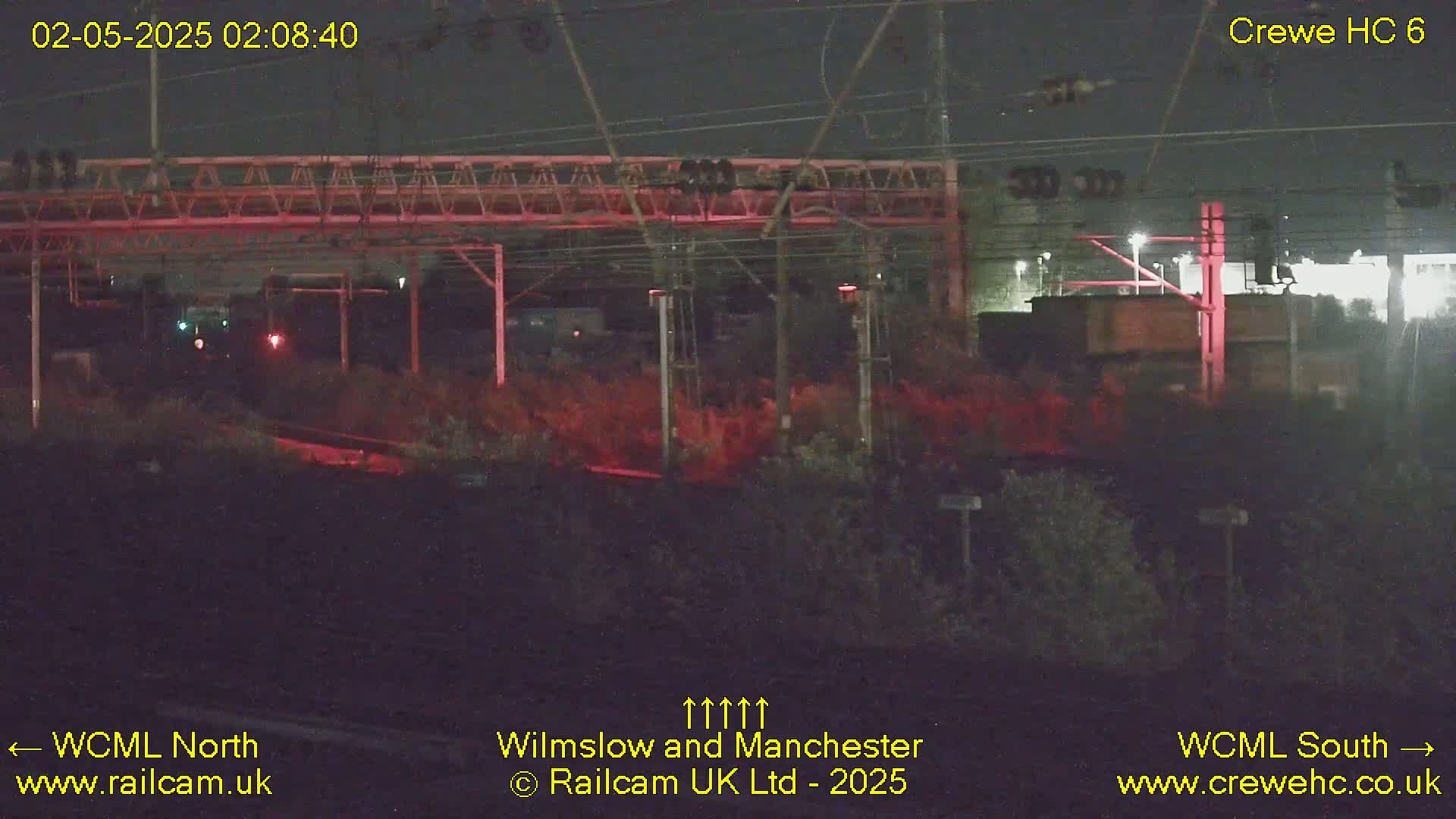 A nighttime view of a railway line with overhead wires and signals, under dark skies.