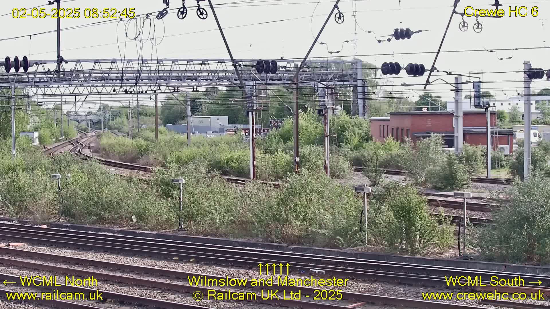 A high-angle view of a railway track network on a partly cloudy day, surrounded by overgrown vegetation and industrial buildings.