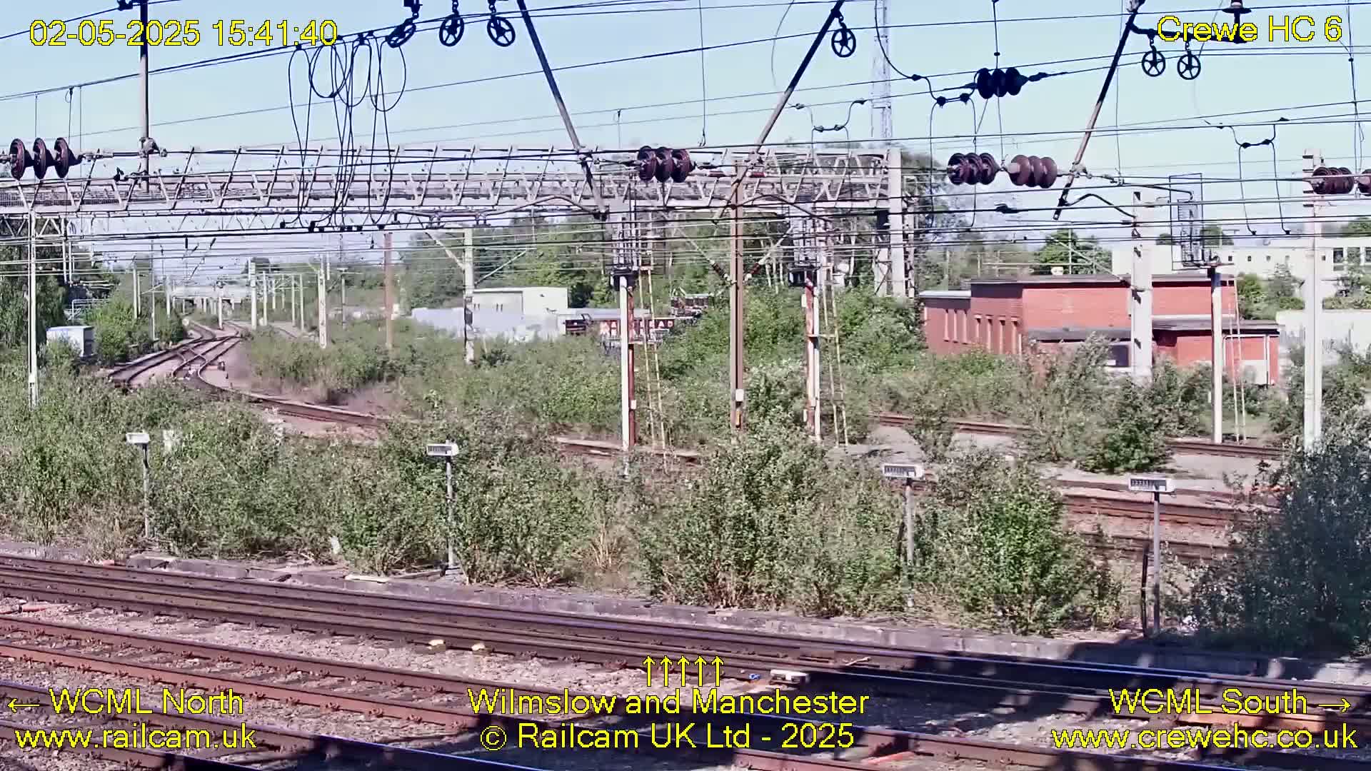 A sunny view of railway tracks and infrastructure, including overhead wires and a brick building, partially obscured by overgrown vegetation.