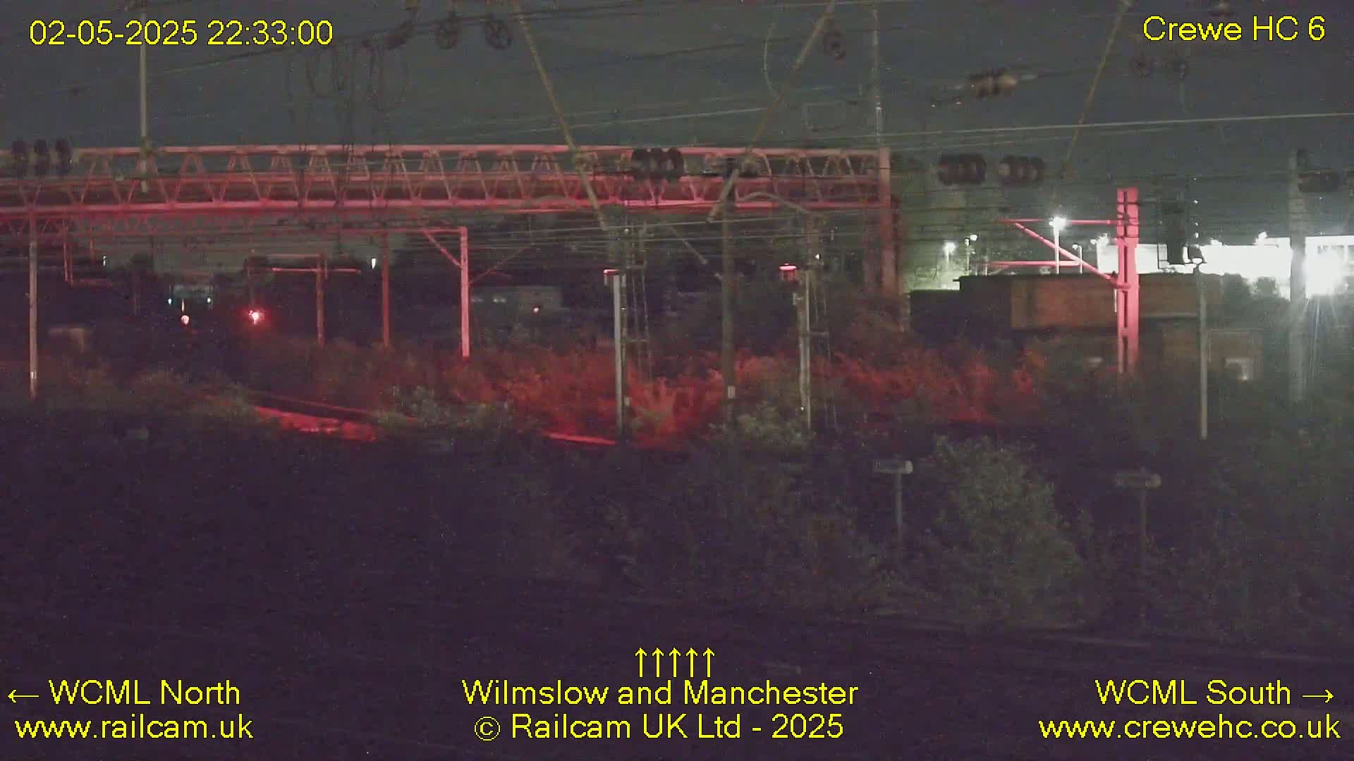 A nighttime view of a railway track with overhead wires and vegetation, illuminated by red lights.