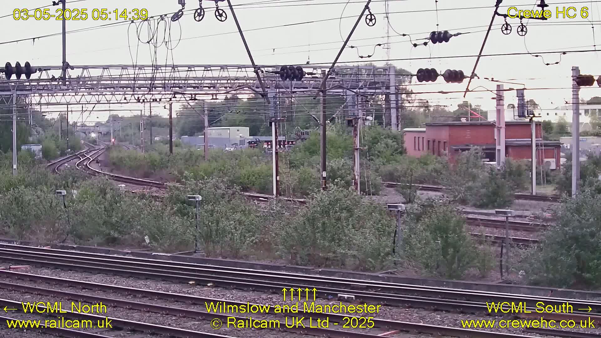 Overgrown railway tracks curve into the distance under an overcast sky, past a low brick building and overhead electrical equipment.
