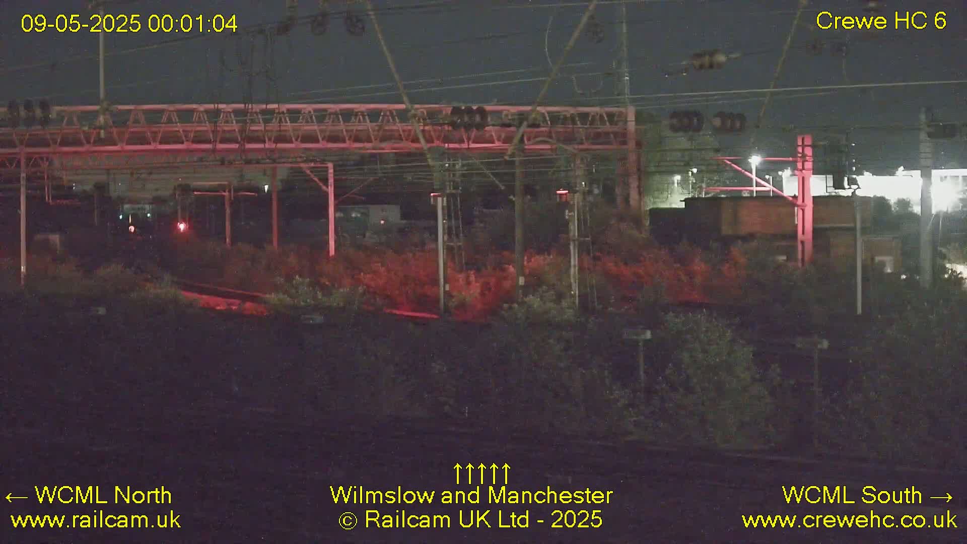 A nighttime view of a train yard with overhead wires and vegetation, illuminated by red lights.