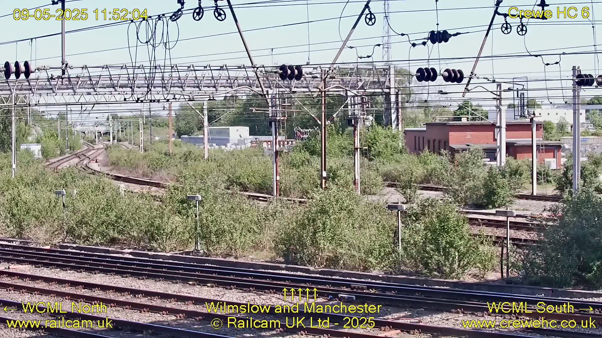 A sunny view of railway tracks, overhead lines, and vegetation surrounding a railway station building.