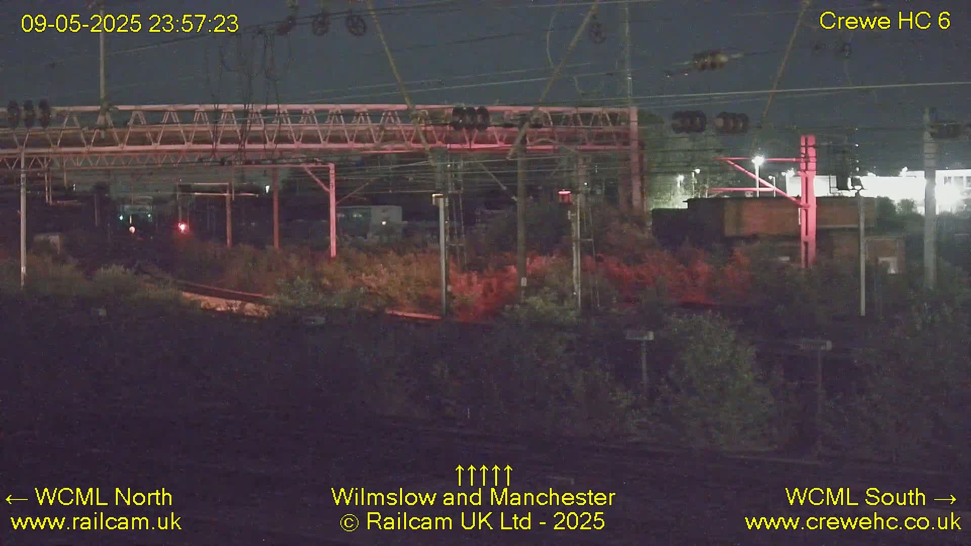A nighttime view of a railway line, with overhead wires and vegetation visible under a dark sky.