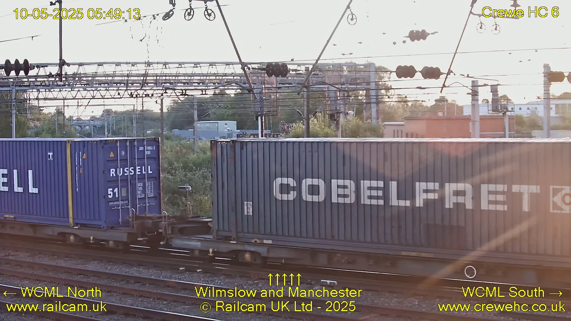 A blue shipping container marked "COBELFRET" on a train passes under overhead wires during the daytime.