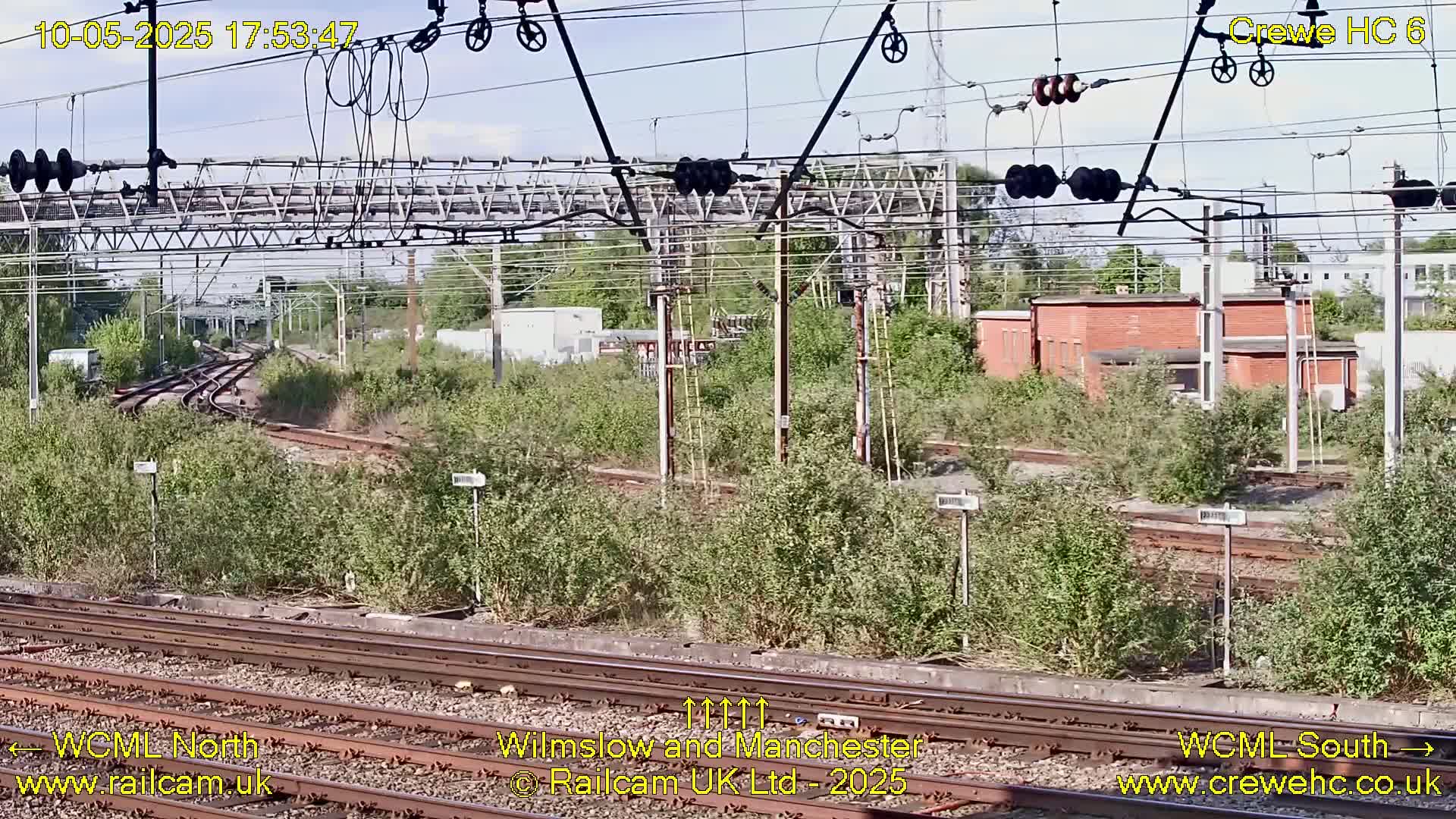A view of railway tracks and infrastructure on a partly sunny day, with overgrown vegetation between the tracks and nearby buildings.