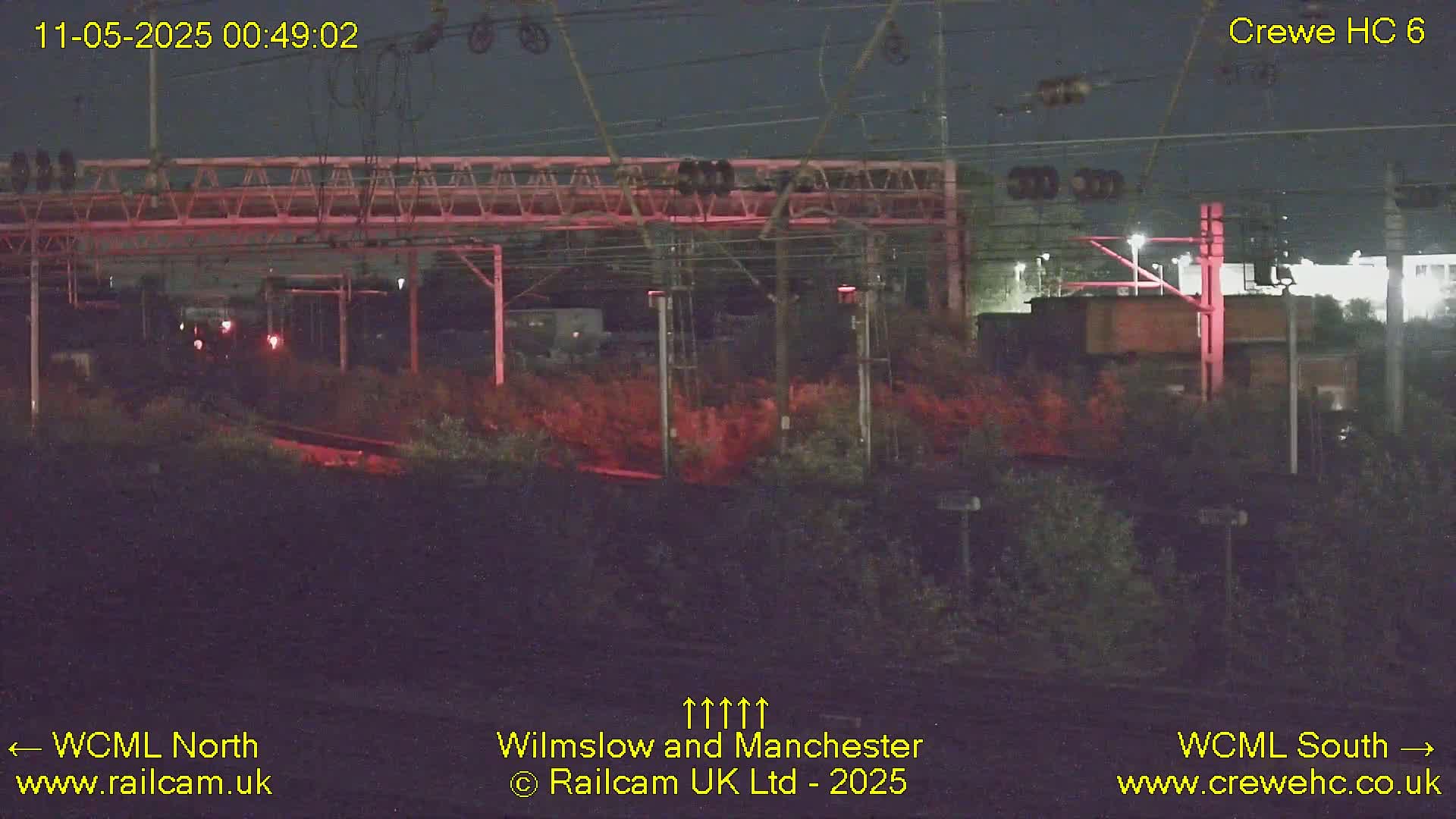 A nighttime view of a railway line with overhead wires and signals, under a dark sky.