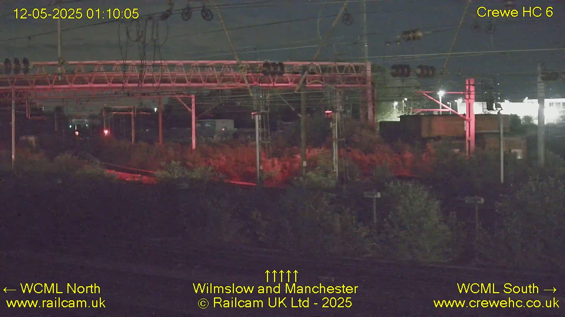 A nighttime view of a railway line with overhead wires, signals, and vegetation.