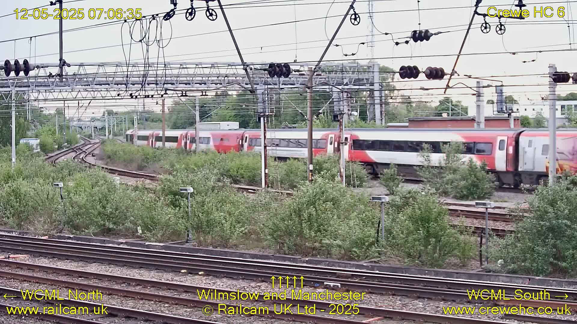 Two red and white passenger trains pass on a railway line amid overcast skies and overgrown vegetation.