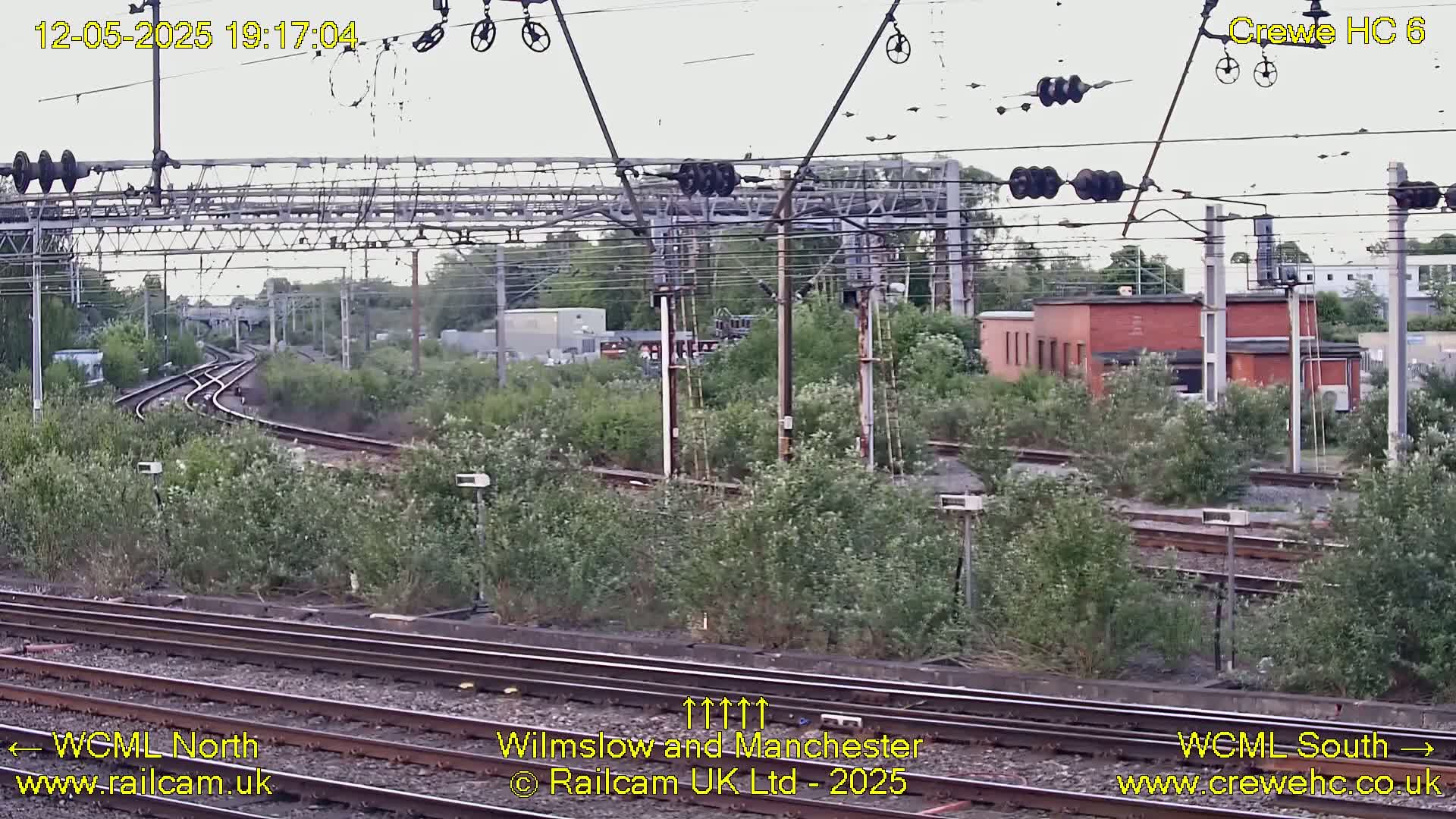 A view of railway tracks and infrastructure on an overcast day, with lush green vegetation alongside the tracks and a brick building in the distance.