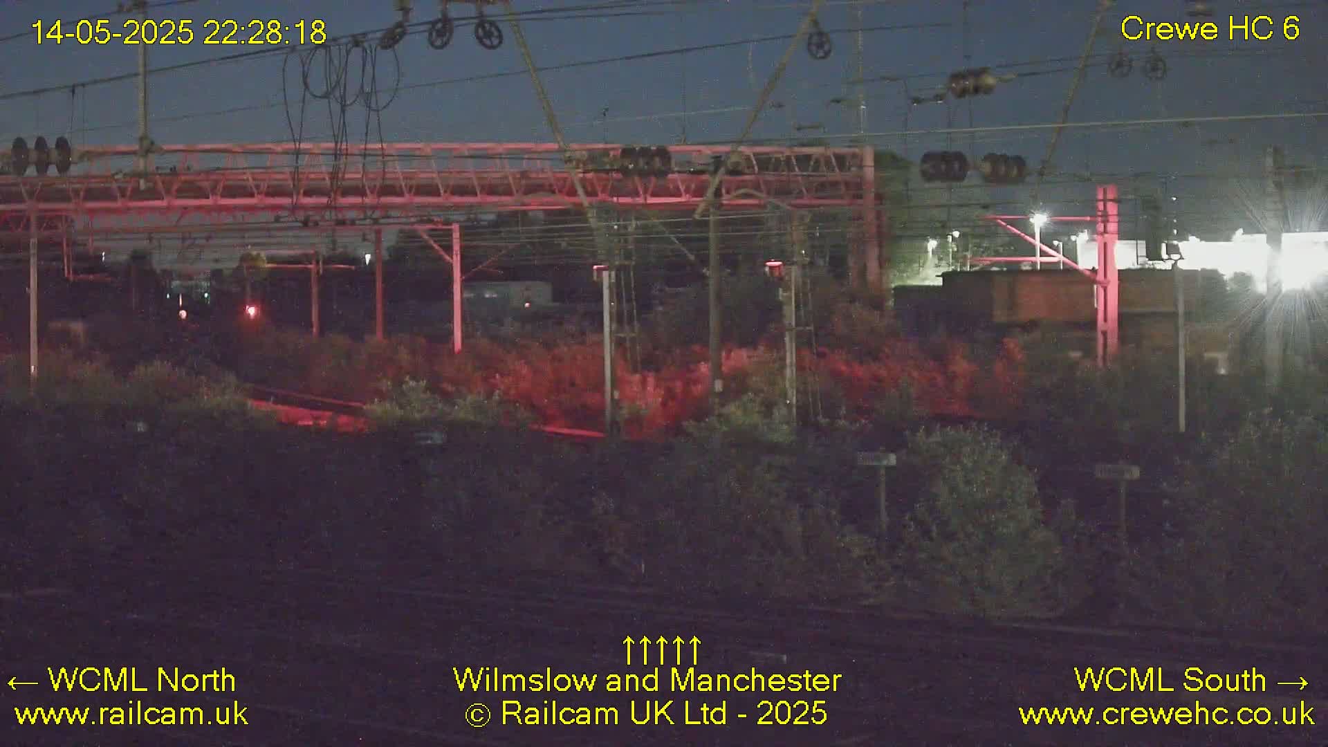 A nighttime view of a railway station, showing tracks, overhead wires, and vegetation, under a dark sky.