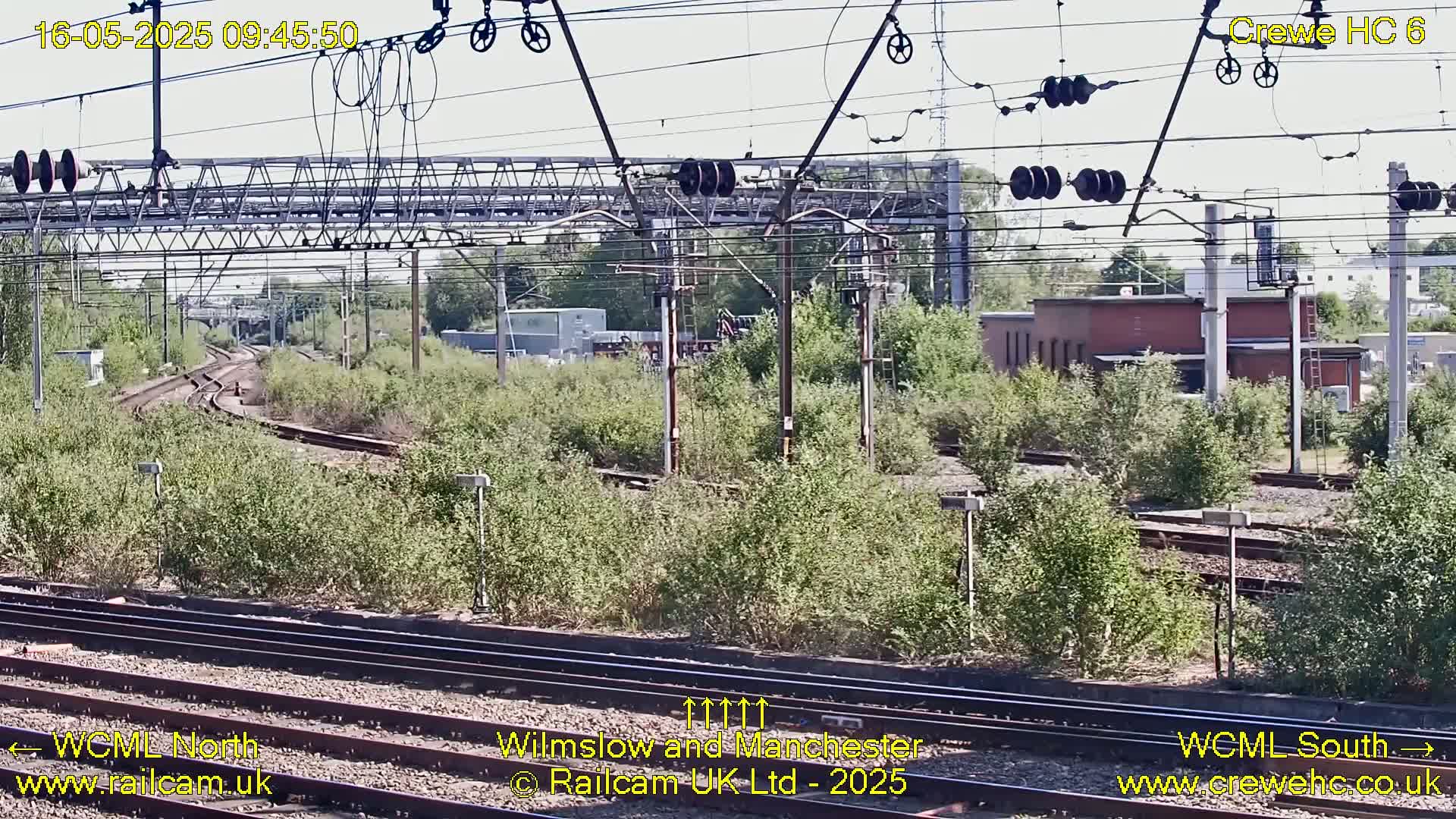 A sunny view of a railway track with overhead lines, branching out into a curving track, surrounded by green vegetation and industrial buildings in the distance.