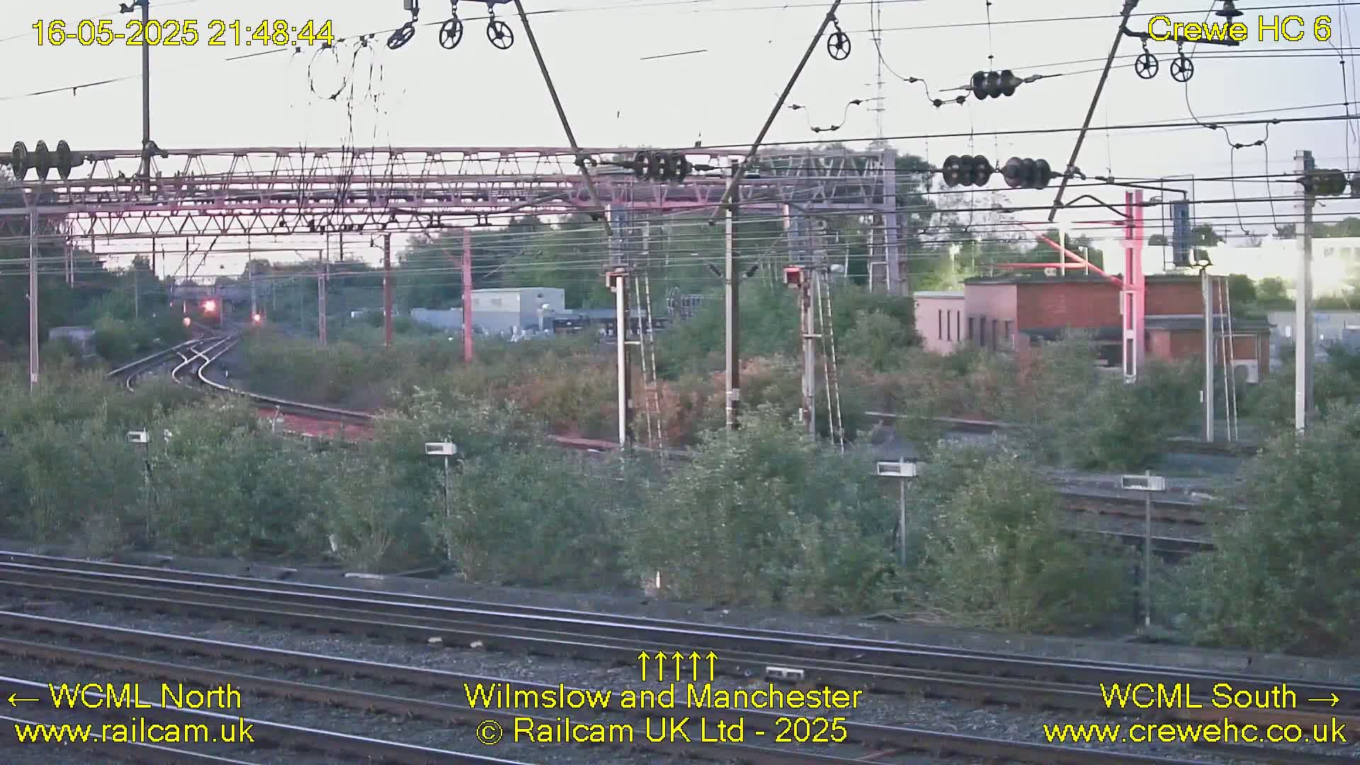 A train approaches a railway junction at dusk, surrounded by lush green vegetation under a partly cloudy sky.