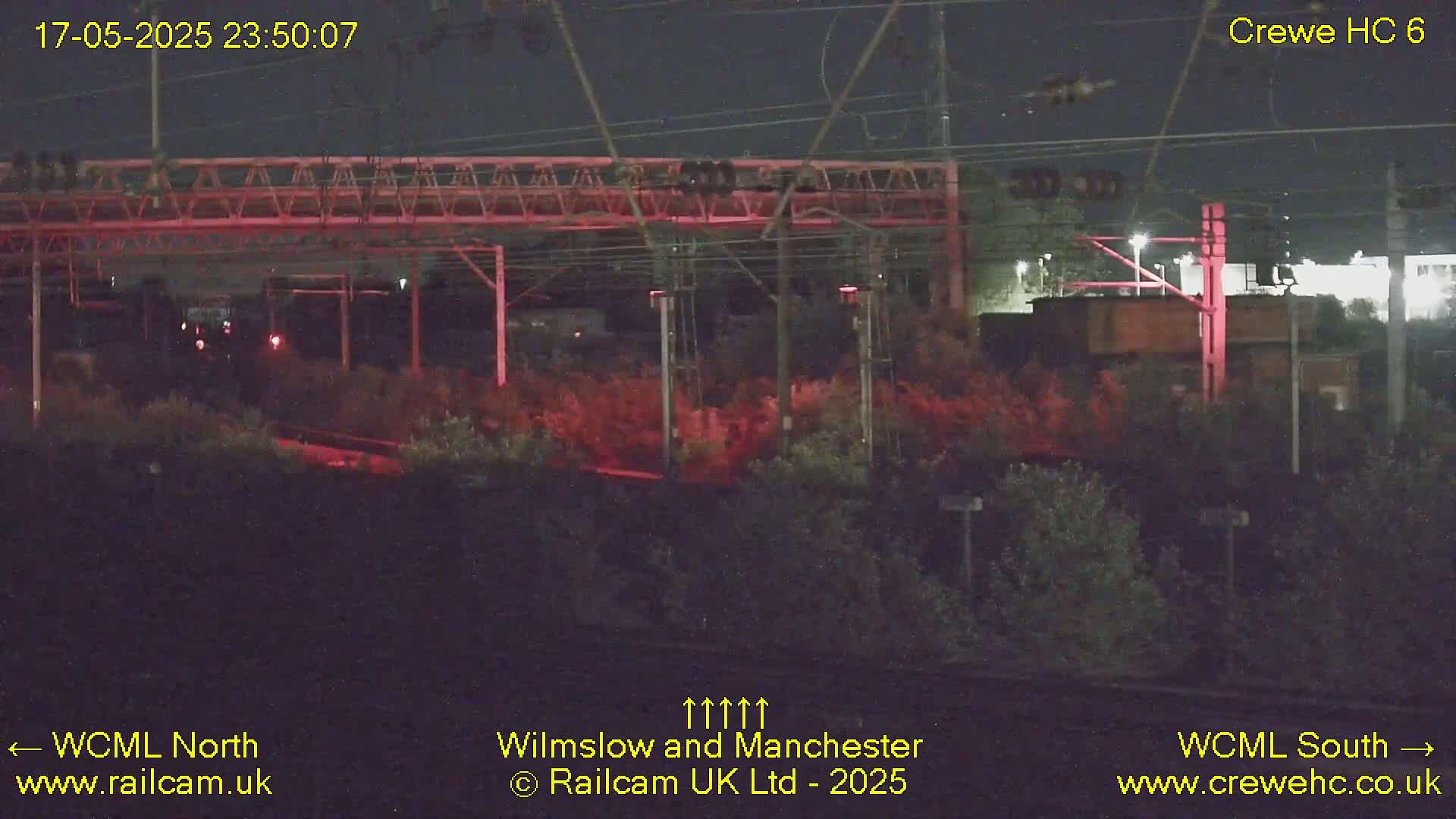 A nighttime view of a railway line with overhead wires, illuminated by lights, shows dark vegetation and a distant building.