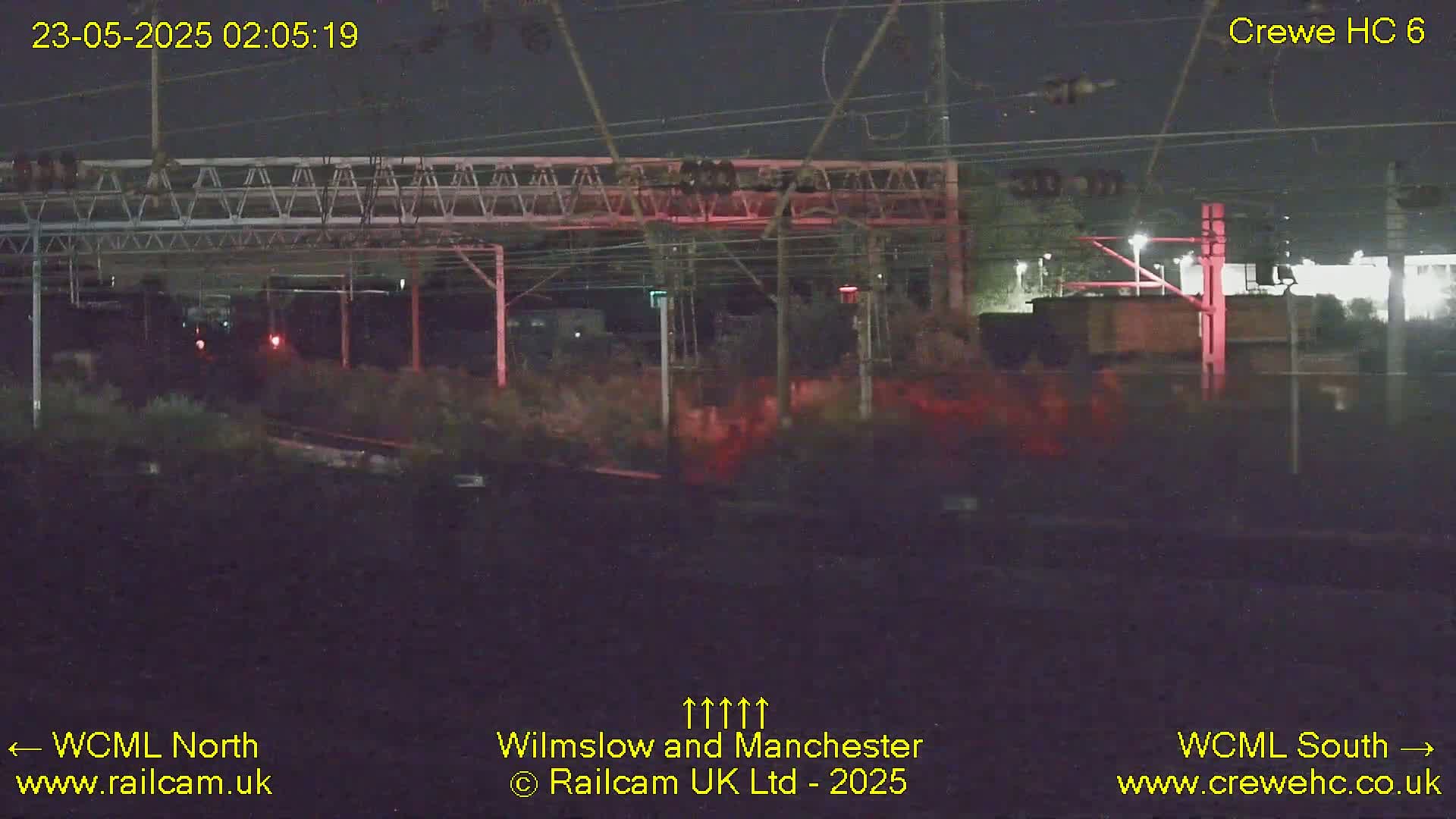 A nighttime view of a railway track with overhead wires and signals, under dark skies.