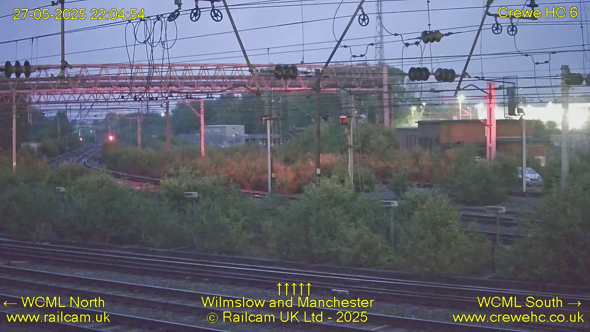 A twilight view of railway tracks, overhead wires, and vegetation, with a distant building visible.