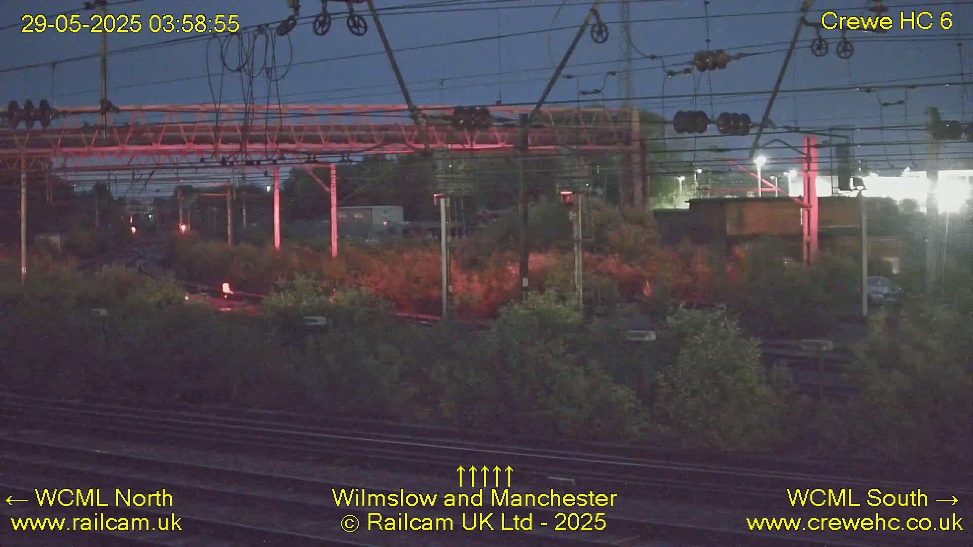 A nighttime view of a railway track, with overhead power lines and surrounding vegetation under a dark sky.