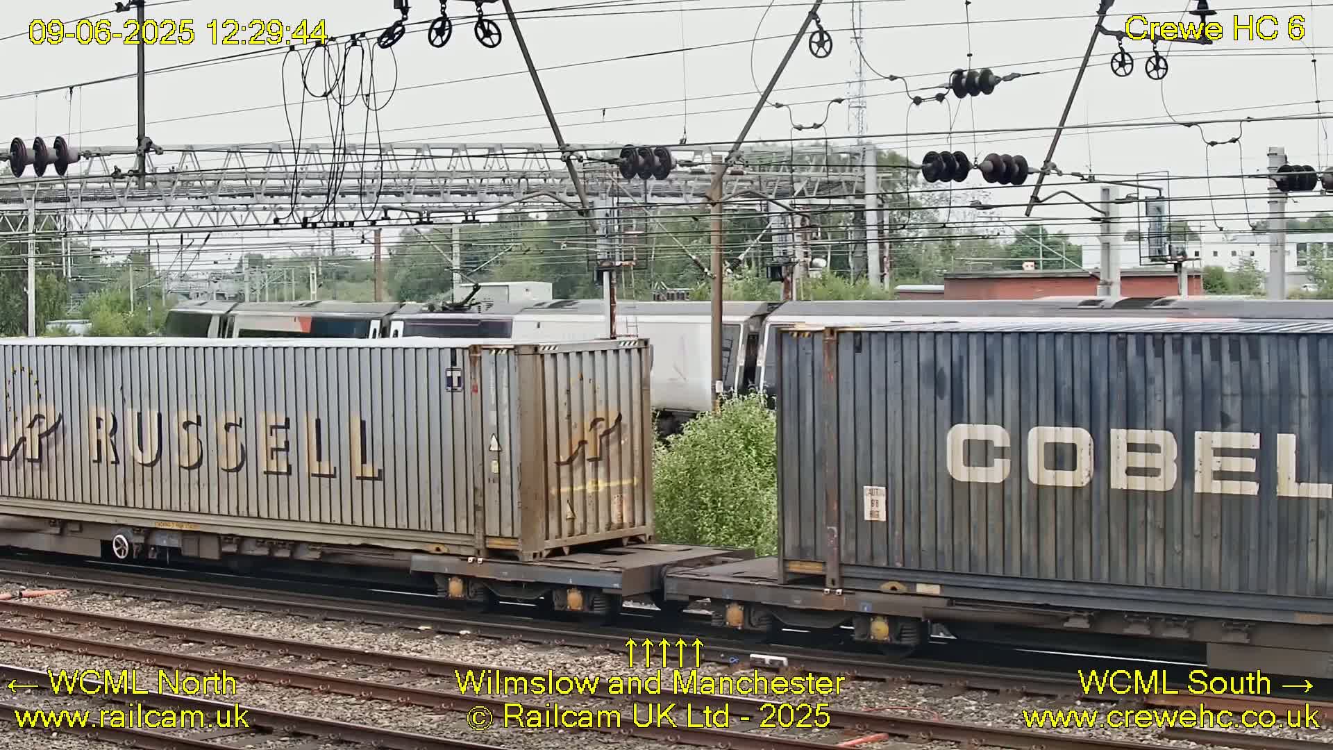 Two shipping containers on a flatcar are positioned on train tracks under an overcast sky, with another train visible in the background.