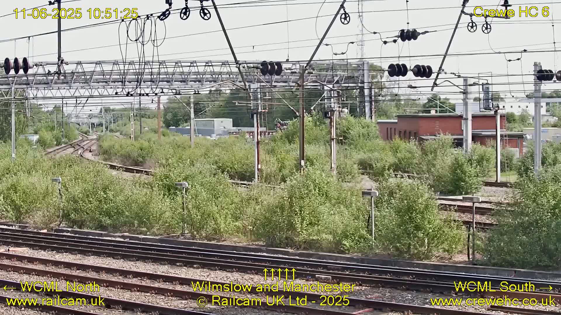 A view of railway tracks and infrastructure on a partly sunny day, with lush green vegetation growing alongside the tracks.