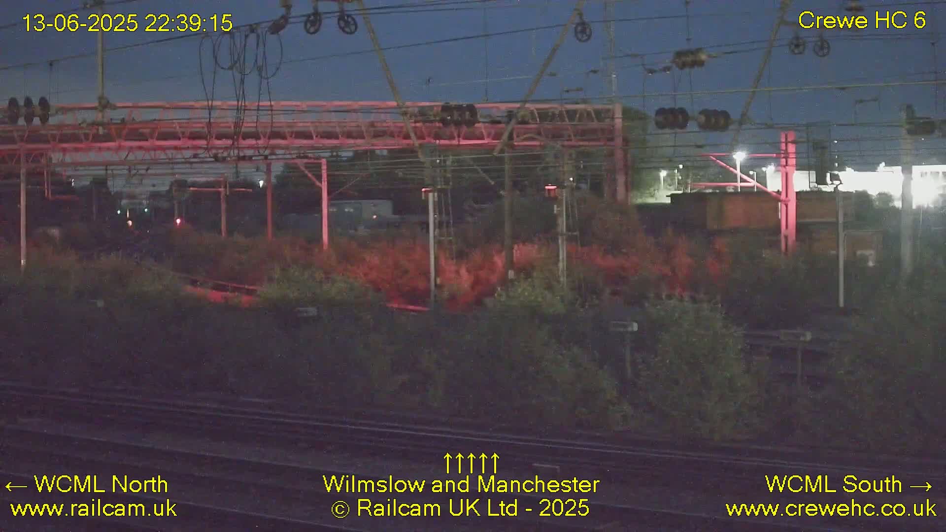 A nighttime view of a railway yard with overhead lines and vegetation, under a dark sky.